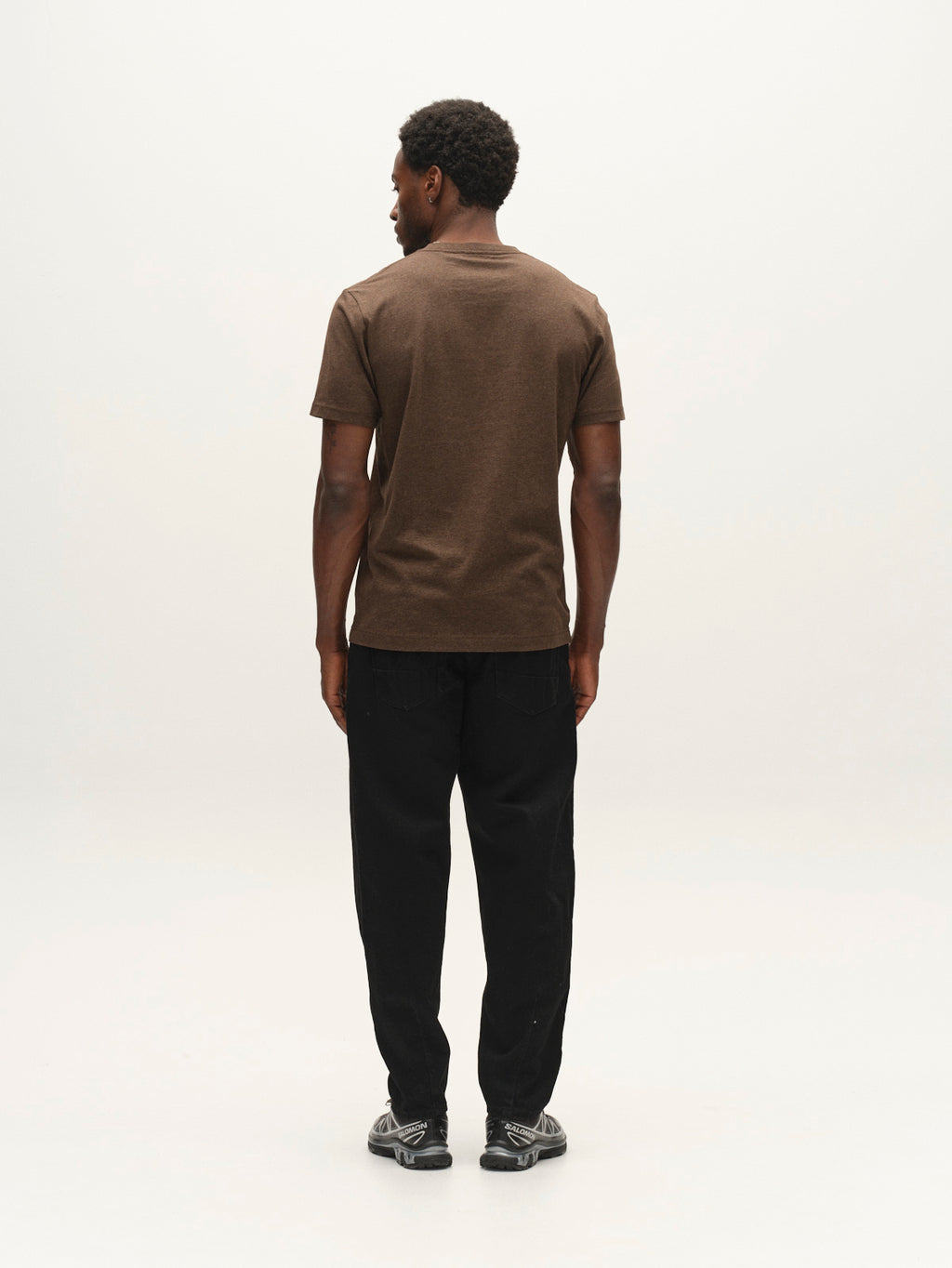 A person stands facing away from the camera, wearing the Gabba GABdune Melange New SS T-Shirt in Brown Slate—its soft melange jersey and subtle chest logo add comfort and texture—paired with black pants and sneakers against a plain, light background.