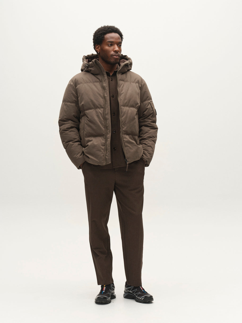 A man wears a brown GABjulien Calle Outerwear - Brown Slate hooded padded insulation jacket over a button-up shirt and dark trousers.