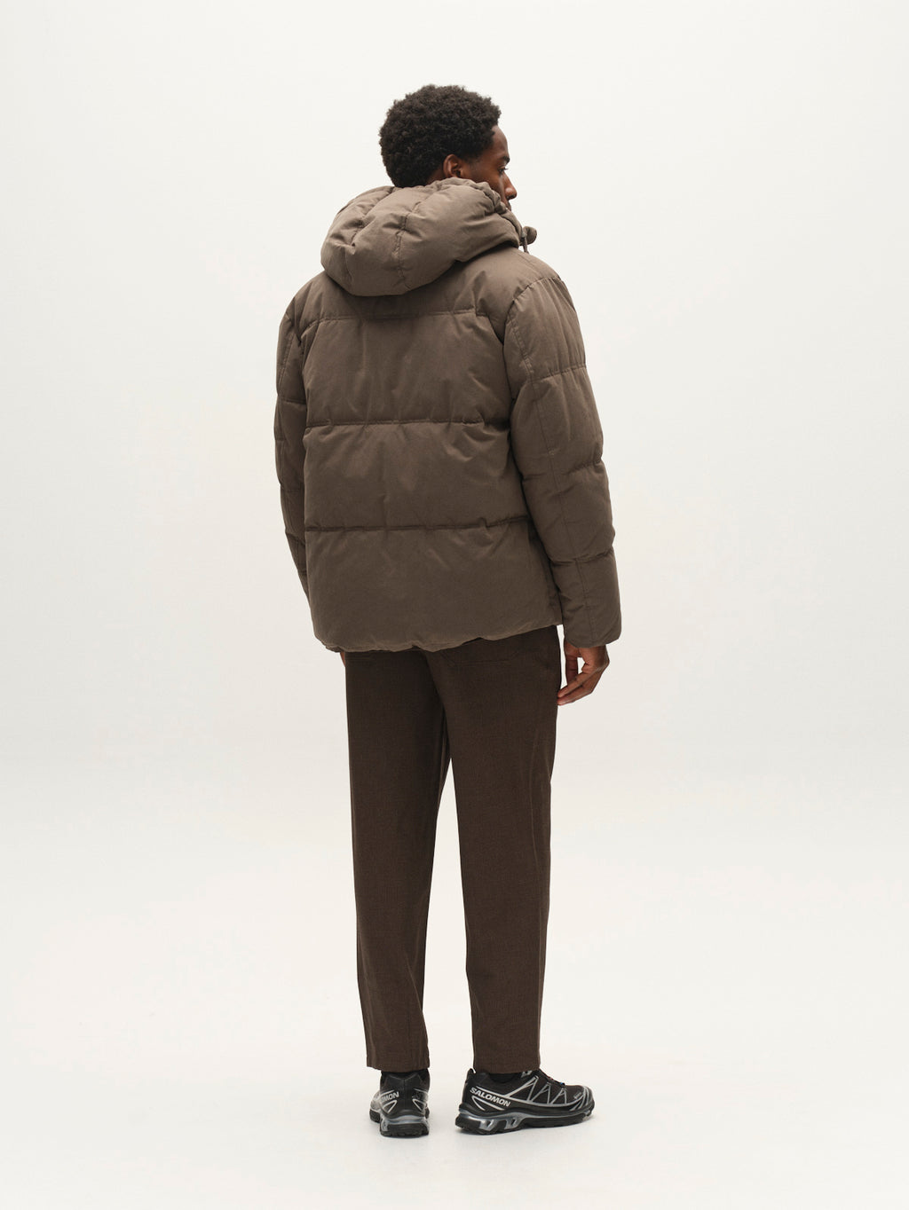A person viewed from behind, wearing a brown GABjulien Calle Outerwear - Brown Slate padded insulation jacket with an adjustable hood, dark trousers, and black sneakers.