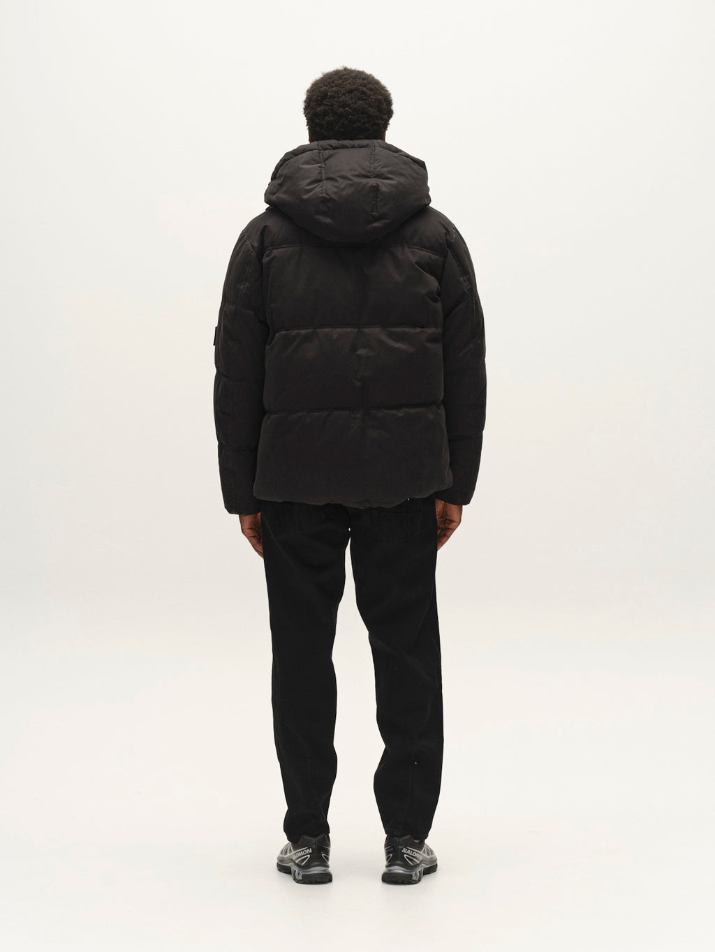 Back view of a person wearing a black GABjulien Calle Outerwear - Black padded insulation jacket with an adjustable hood, black trousers, and athletic sneakers.
