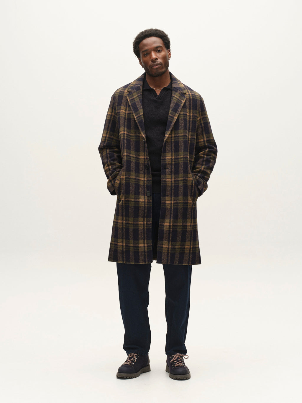 Wearing the Gabba GABleeds Chezy Coat in Forest Night, a man stands against a plain background with his hands in his coat pockets. The long, wool-blend checked coat—a true outerwear essential—features a tailored silhouette that adds refined structure to his ensemble of a black shirt and dark pants.