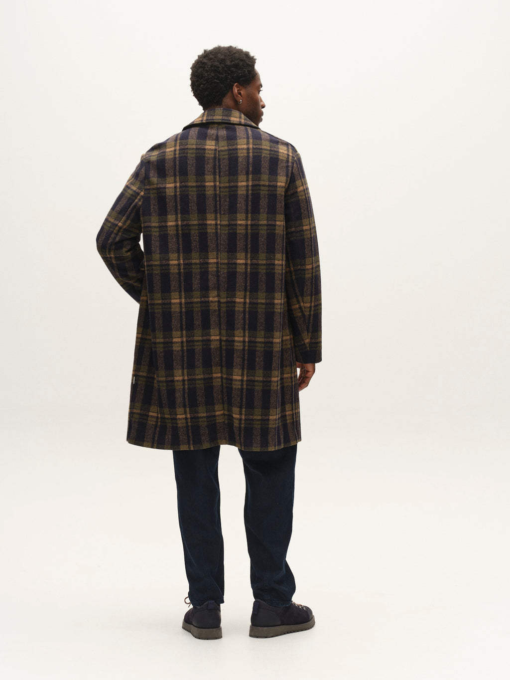 A person stands facing away, wearing the Gabba GABleeds Chezy Coat in Forest Night—a wool-blend checked coat with a tailored silhouette—paired with dark pants and dark shoes against a plain, light background.