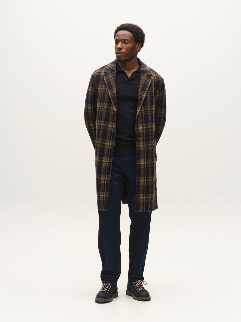 A man stands facing forward, wearing the Gabba GABleeds Chezy Coat in Forest Night, a wool-blend checked coat with a tailored silhouette, layered over a dark shirt and navy pants with dark shoes. His hands are placed behind his back as he gazes slightly to the side against a plain light background. This outerwear essential from Gabba adds timeless style to his look.
