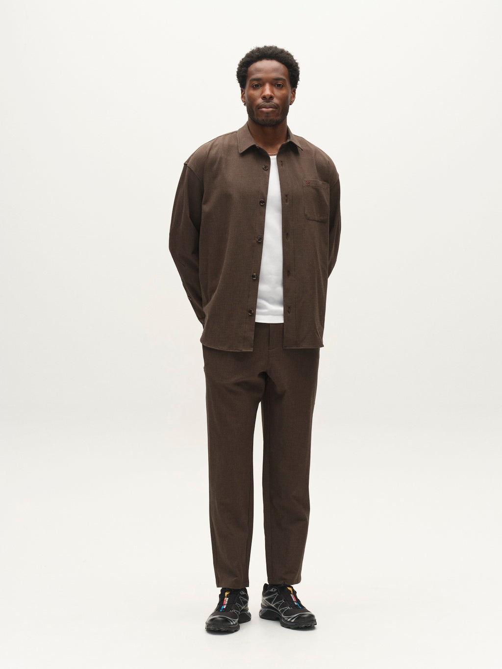 A man stands against a plain white background, wearing the Gabba GABjens Over Rubin LS Overshirt in Brown Slate with matching brown pants, a white t-shirt, and black sneakers. His hands are behind his back as he faces forward.