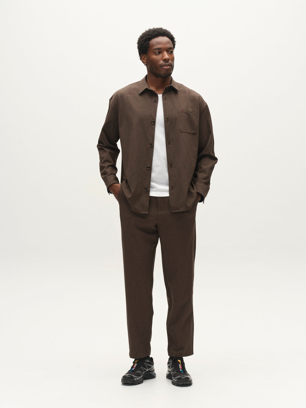 A man stands in a neutral studio wearing the Gabba GABjens Over Rubin LS Overshirt in Brown Slate with an oversized fit, matching brown pants, a white t-shirt underneath, and black sneakers. He has his left hand in his pocket and looks slightly to his right.