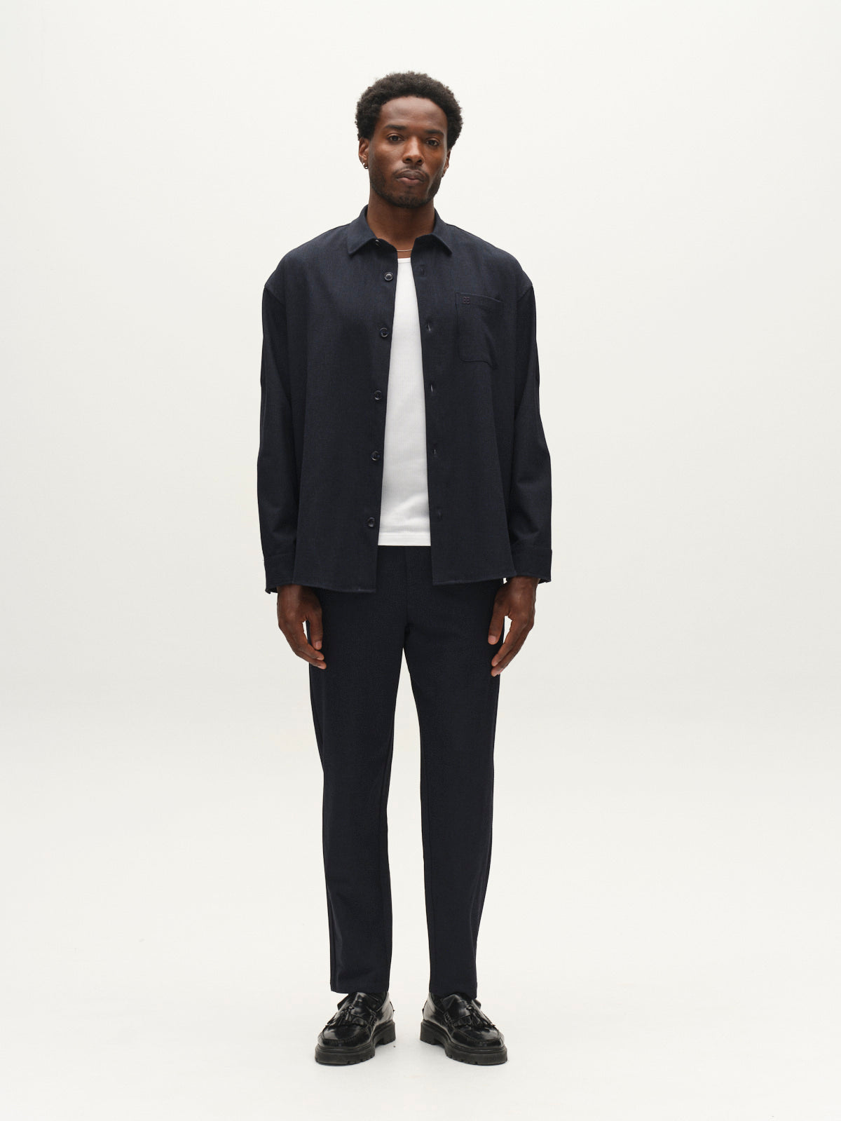 A man stands facing forward against a plain white background, wearing the Gabba GABjens Over Rubin LS Overshirt in Blue Black over a white t-shirt, paired with matching navy pants and black shoes.