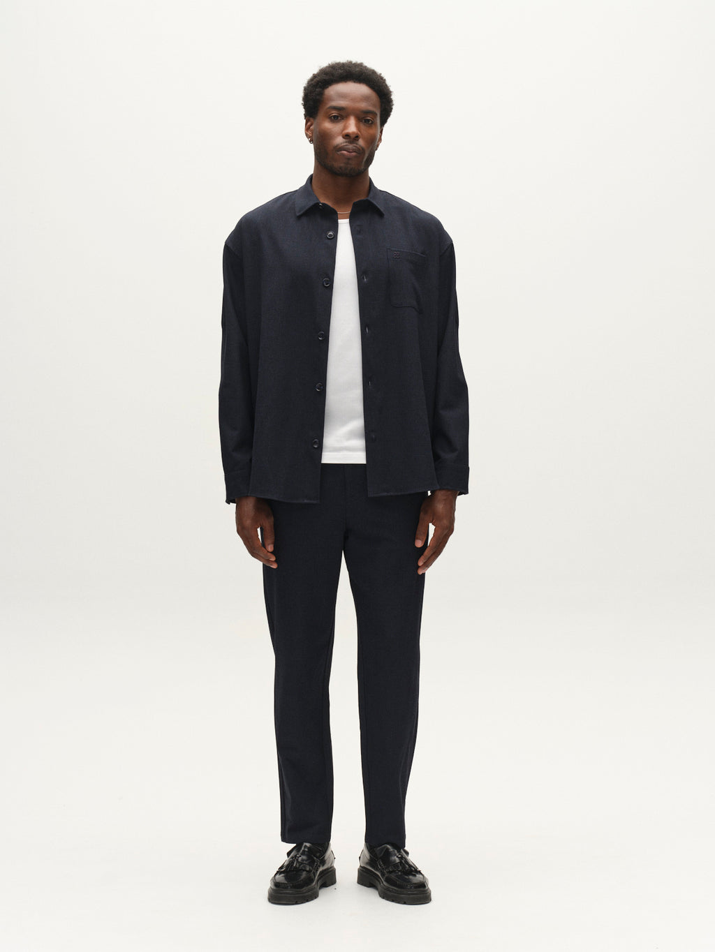 A man stands facing forward against a plain white background, wearing the Gabba GABjens Over Rubin LS Overshirt in Blue Black over a white t-shirt, paired with matching navy pants and black shoes.