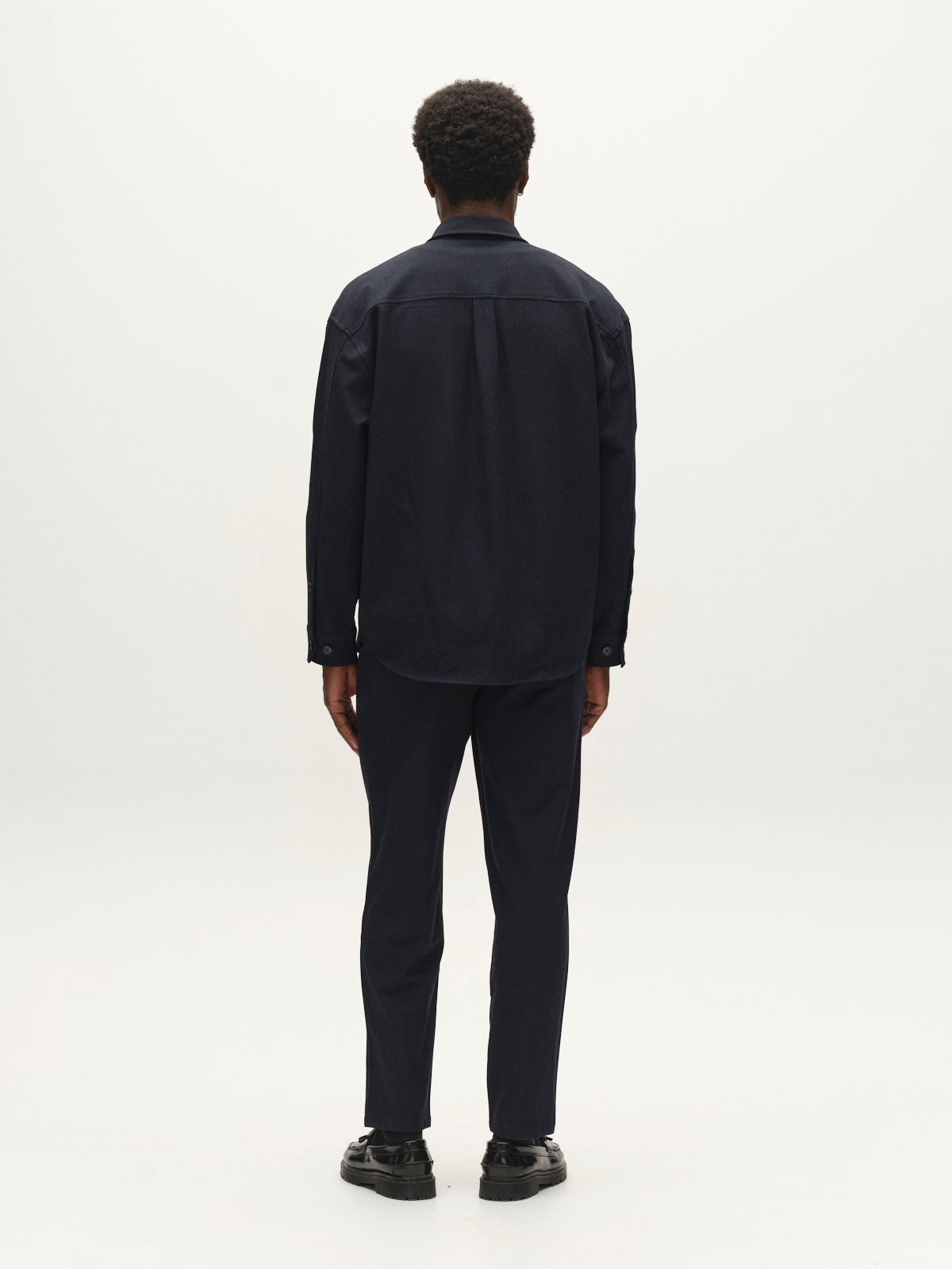 A person with short curly hair, dressed in the Gabba GABjens Over Rubin LS Overshirt in Blue Black and dark stretch dobby pants, stands facing away from the camera against a plain white background.