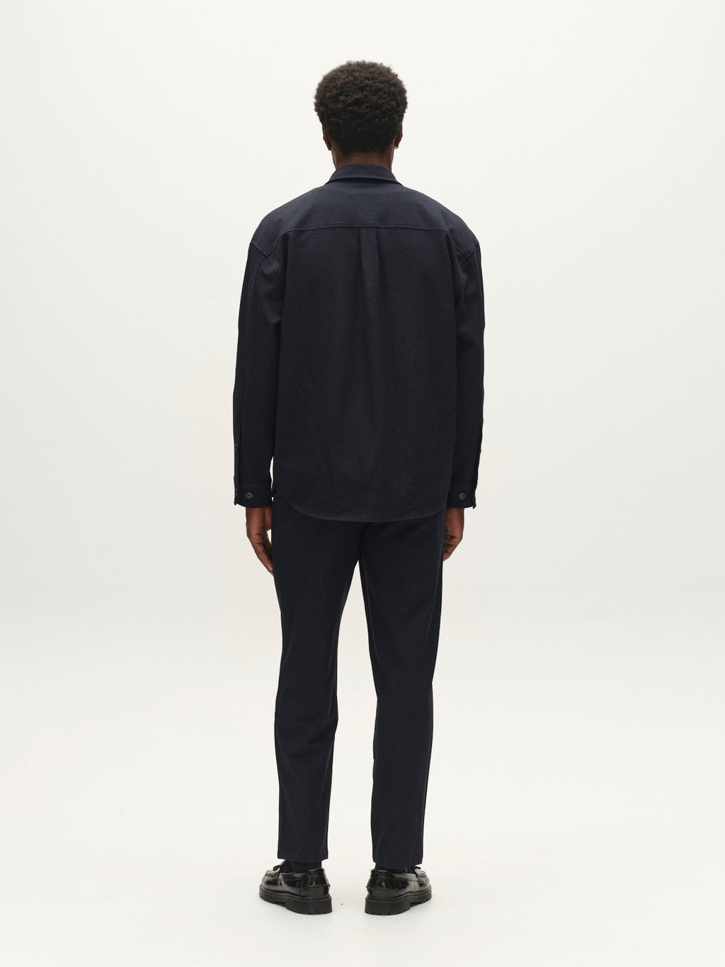 A person with short curly hair, dressed in the Gabba GABjens Over Rubin LS Overshirt in Blue Black and dark stretch dobby pants, stands facing away from the camera against a plain white background.