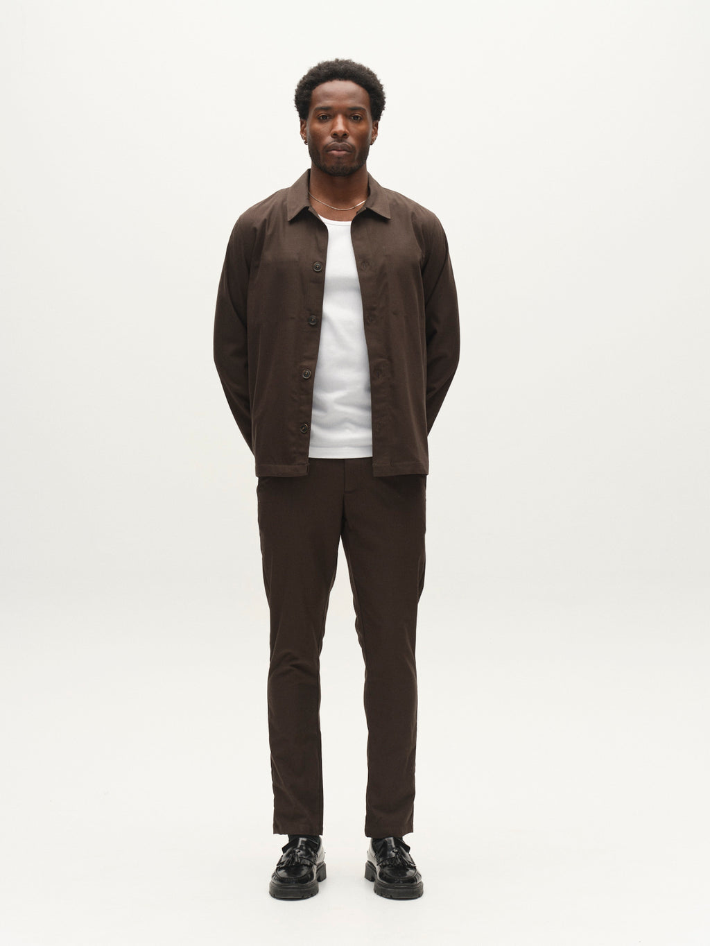 A man stands facing forward against a plain white background, wearing the Gabba GABtogger Base Deme LS Overshirt in Forest Night—a regular fit button-up jacket crafted from micro-pattern woven fabric—paired with matching brown pants, a white shirt, and black shoes.