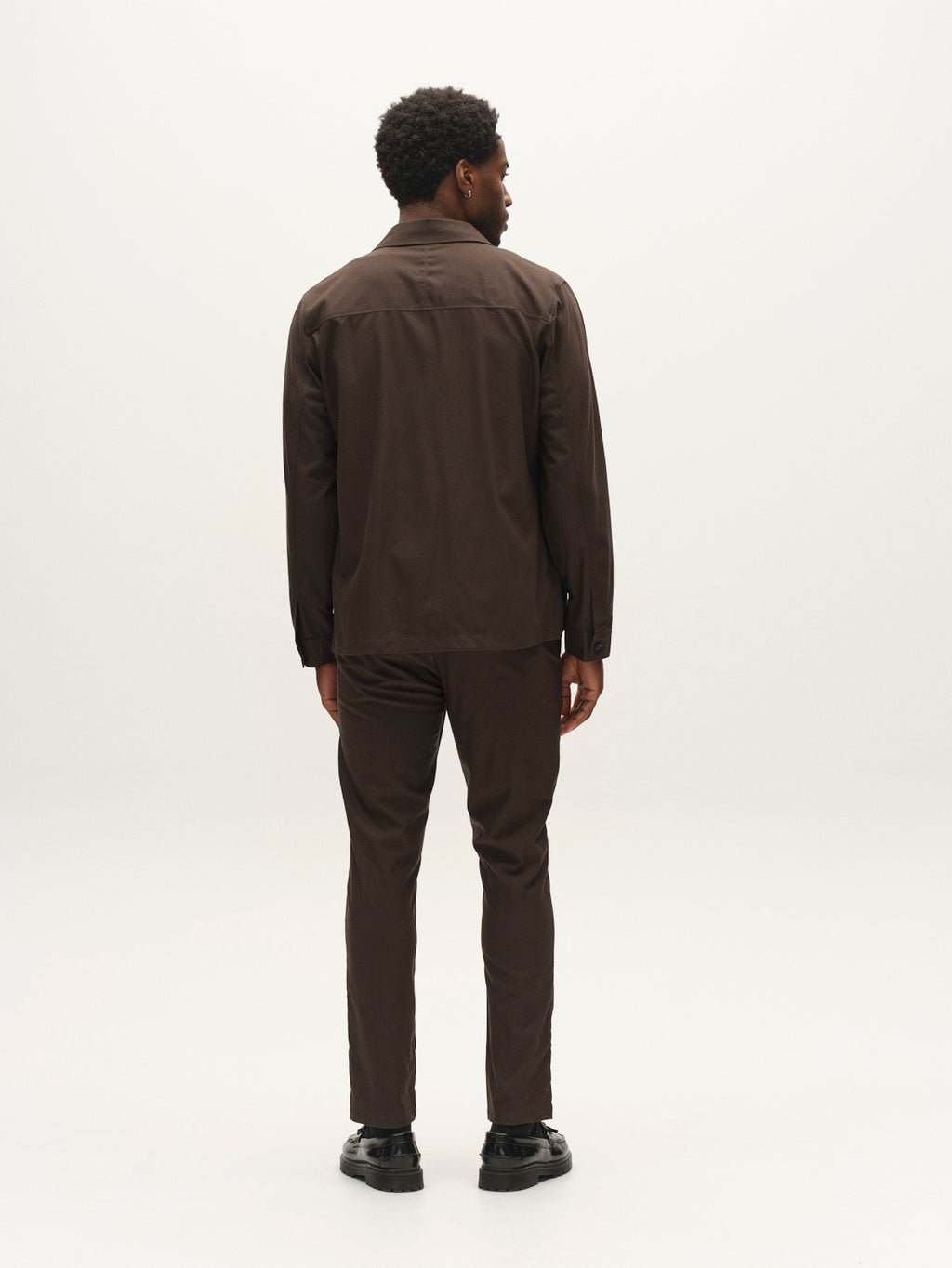 A man stands facing away, wearing the Gabba GABtogger Base Deme LS Overshirt in Forest Night—a dark brown lightweight piece crafted from micro-pattern woven fabric—and matching pants, paired with black shoes against a plain white background.