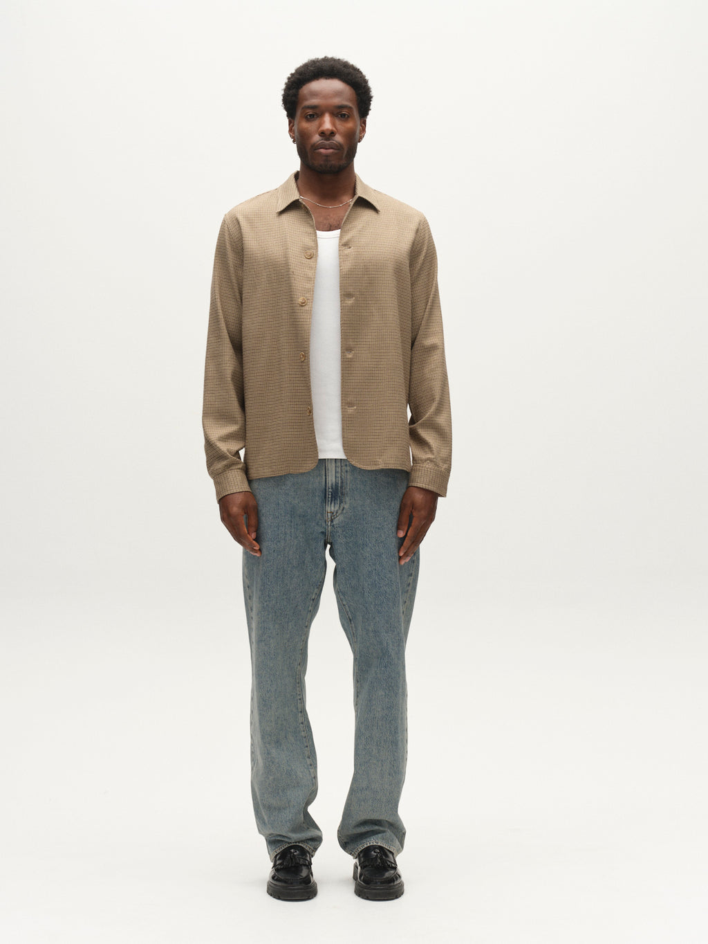 A man stands facing forward wearing the Gabba GABfelix Brit LS Shirt in Dark Sand—a relaxed fit tan button-up—layered over a white t-shirt, paired with blue jeans and black shoes against a plain white background.