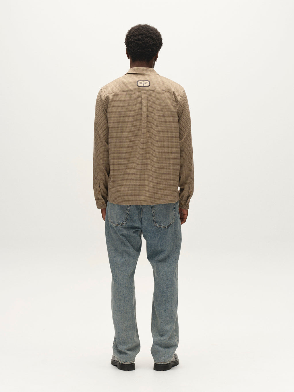 A person with short hair stands facing away, wearing the Gabba GABfelix Brit LS Shirt in Dark Sand, a relaxed-fit shirt crafted from lightweight woven fabric, paired with loose blue jeans, against a plain light background.