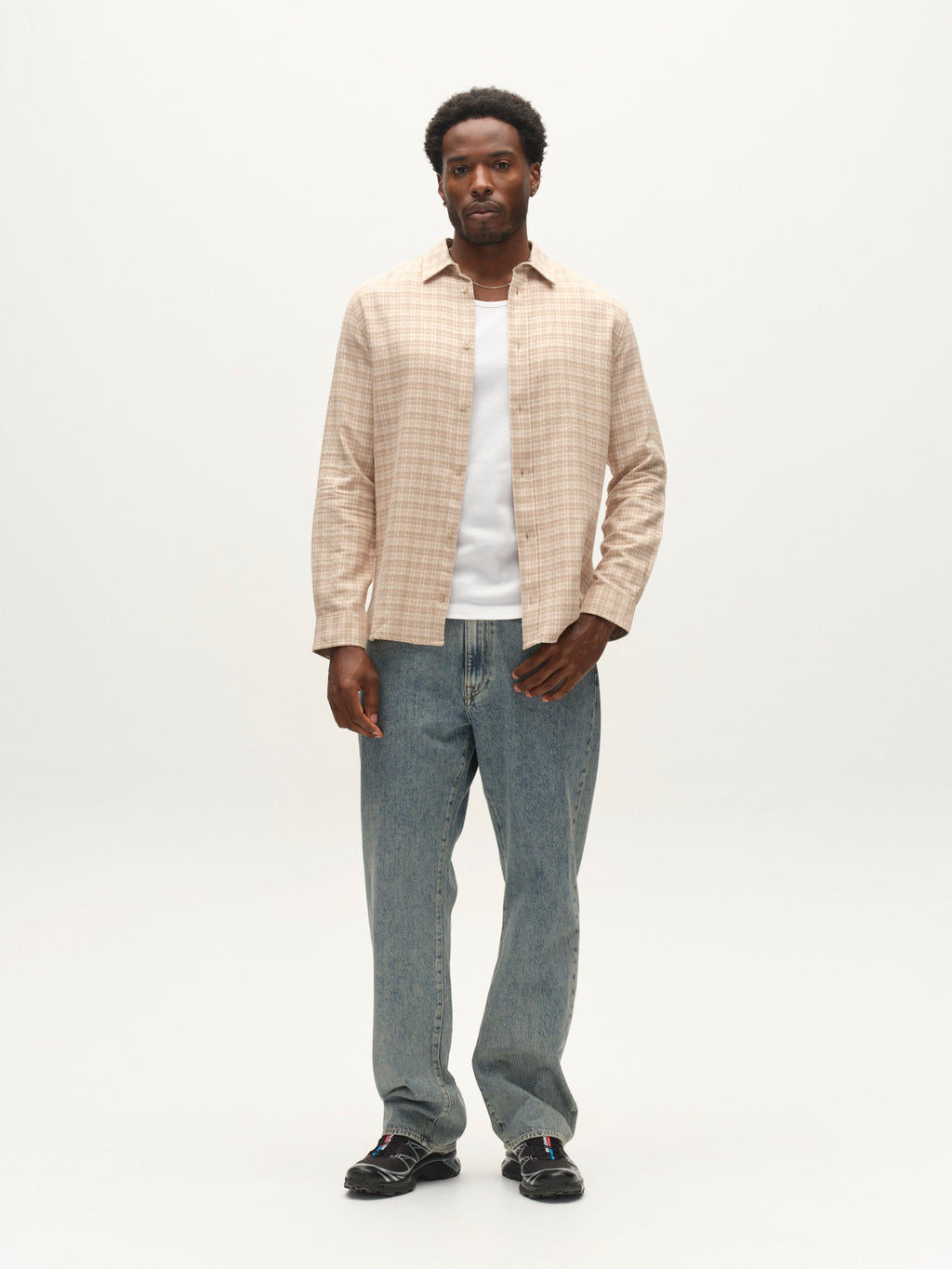 A man stands against a plain white background wearing the Gabba GABsven Reg TX2 LS Shirt in Sand Crocker over a white t-shirt, paired with blue jeans and black shoes. He has short curly hair and a neutral expression.
