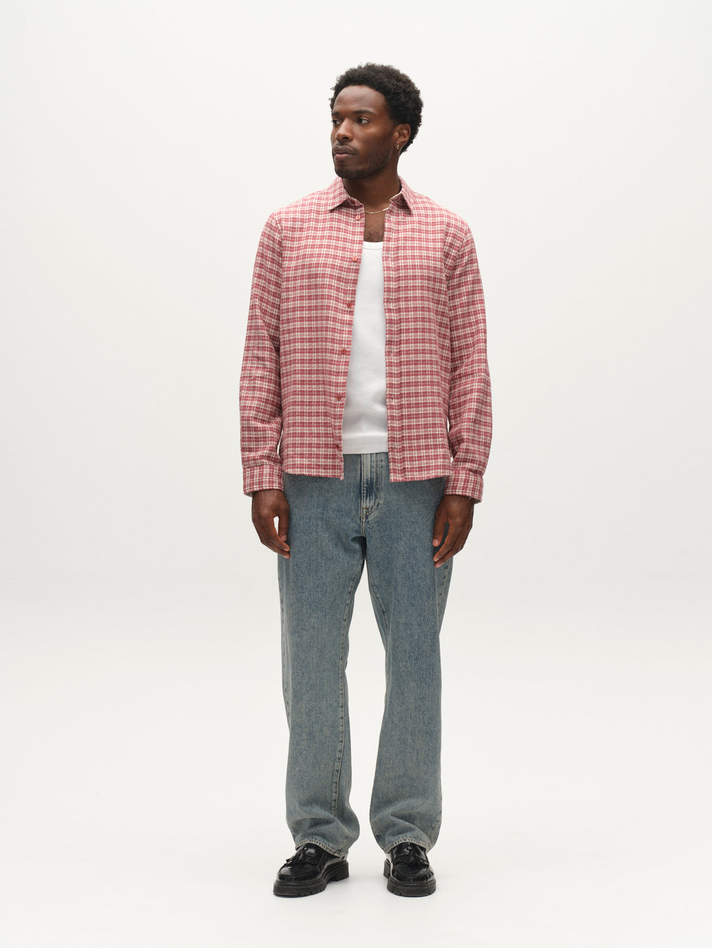 A man stands facing forward against a plain white background, wearing the Gabba GABsven Reg TX2 LS Shirt in Brick Red—a regular-fit, yarn-dyed cotton shirt with a checked texture—layered over a white t-shirt, paired with light blue jeans and black shoes.
