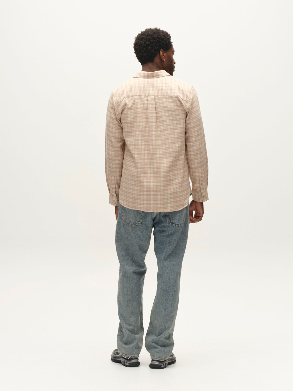 A man stands facing away, wearing the Gabba GABsven Reg TX2 LS Shirt in Sand Crocker, paired with blue jeans and black sneakers, against a plain white background.