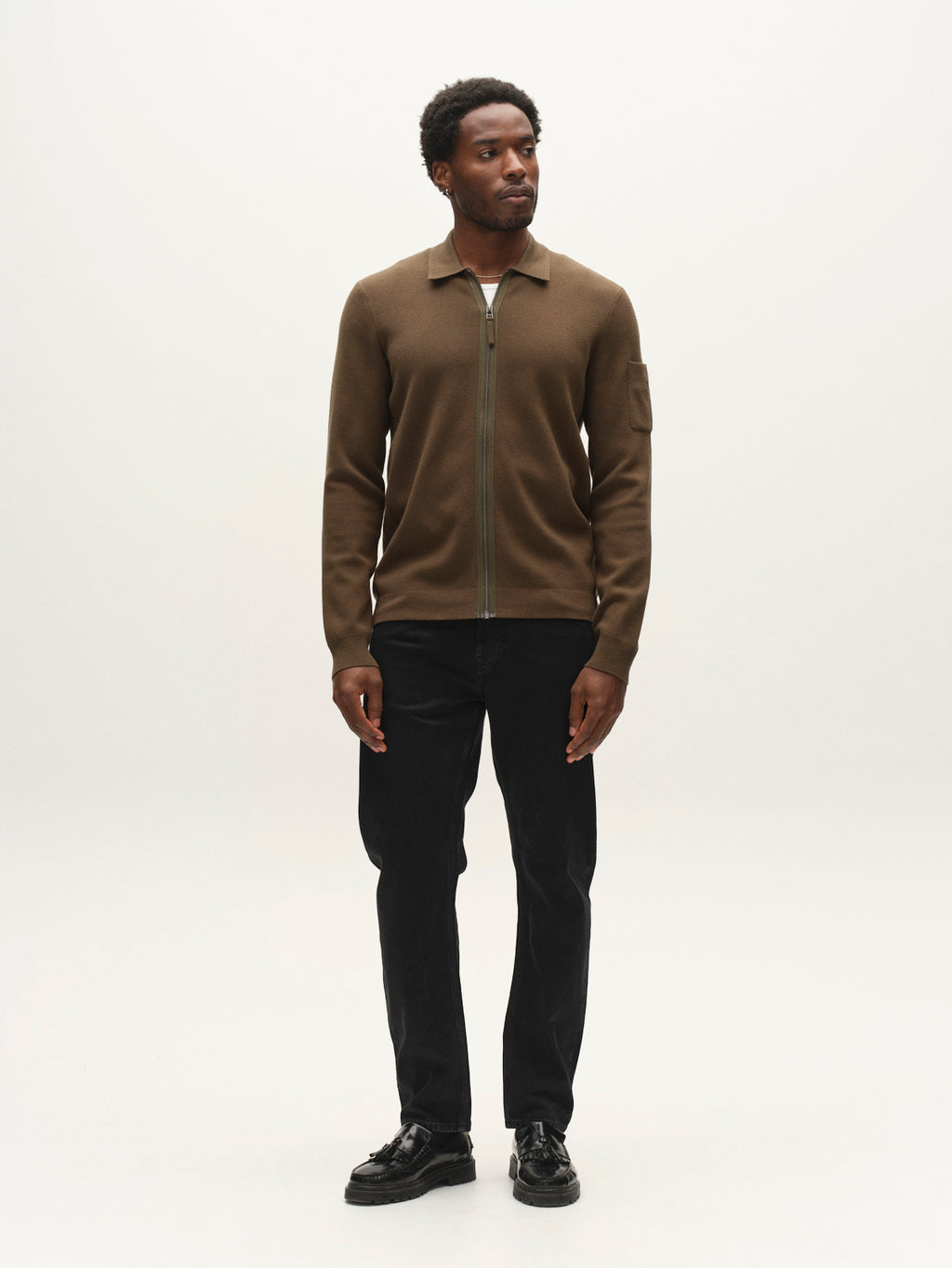 A man stands facing slightly left, wearing the Gabba GABadriel Zip Lume LS Knit in Forest Night—a regular-fit zip sweater—paired with black pants and black shoes against a plain white background.