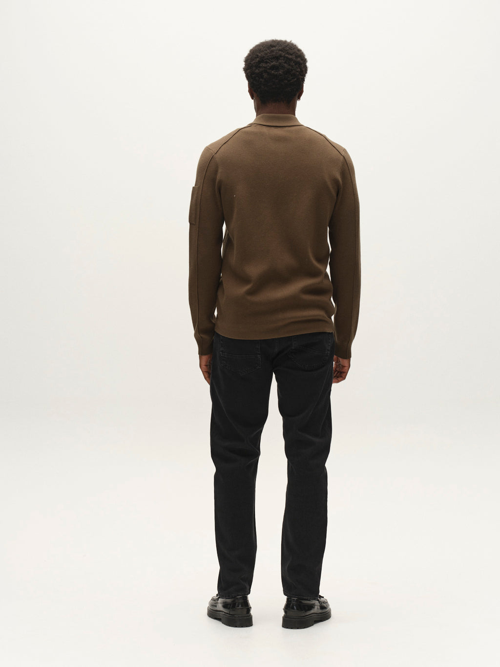 A person with short curly hair, dressed in the Gabba GABadriel Zip Lume LS Knit in Forest Night, along with black jeans and black shoes, stands facing away from the camera against a plain white background.