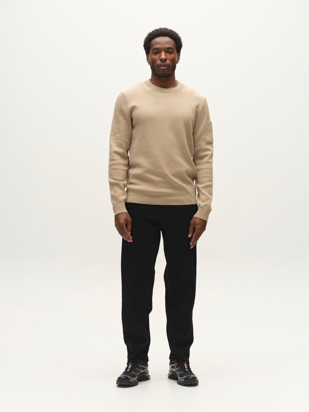A man stands facing forward against a plain white background, wearing the Gabba GABadriel O-Neck Lume LS Knit in Sand Crocker—a regular fit viscose blend sweater—paired with black pants and black sneakers.