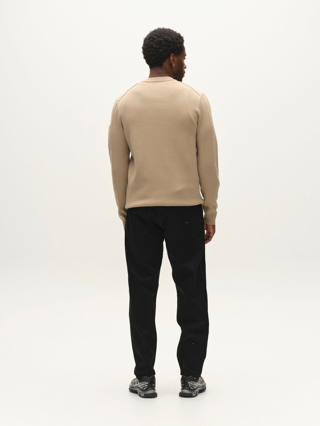 A man stands facing away, wearing the Gabba GABadriel O-Neck Lume LS Knit sweater in Sand Crocker—a regular fit crew neck crafted from a soft viscose blend—paired with black pants and gray athletic shoes, set against a plain white background.