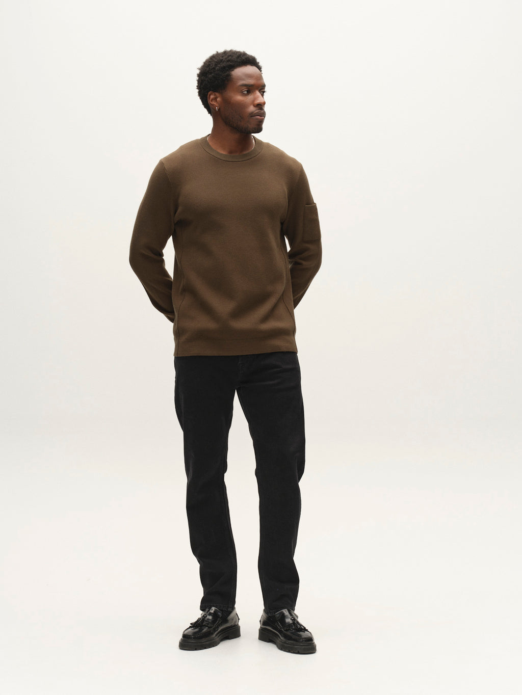 A man stands against a plain white background, wearing the Gabba GABadriel O-Neck Lume LS Knit in Forest Night—a mid-weight viscose-blend crew neck sweatshirt—paired with black pants and black shoes. His hands are behind his back as he looks slightly to the side.