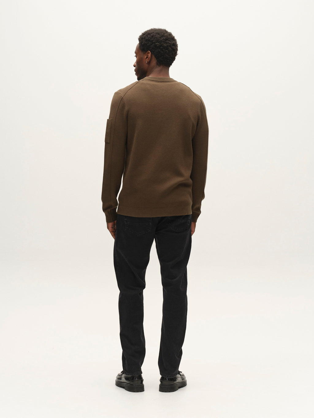 A man stands facing away from the camera, wearing the Gabba GABadriel O-Neck Lume LS Knit sweater in Forest Night, paired with black jeans and black shoes, against a plain light background.