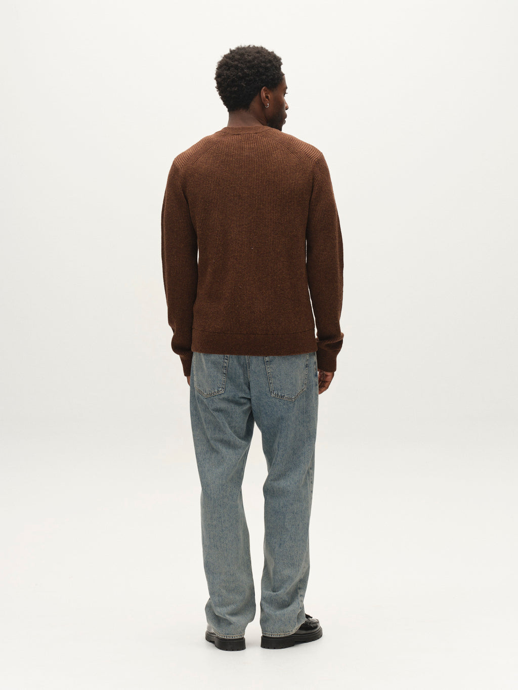 A person stands facing away, wearing the Gabba GABremy Cardi Flash LS Knit in Brown Slate, paired with light blue jeans and black shoes against a plain white background.
