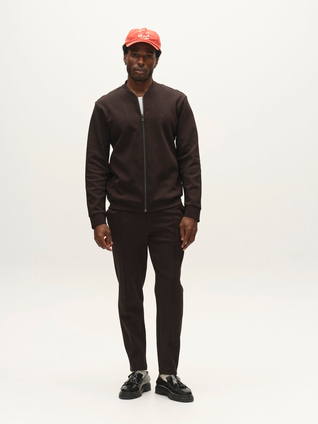 A man stands facing forward on a plain white background, wearing the Gabba GABnik Camp Lock Sweat in Meteorite—a modern zip-up jacket crafted from mid-weight interlock fabric with a sleek bomber collar—paired with matching pants, black shoes, and a red baseball cap.
