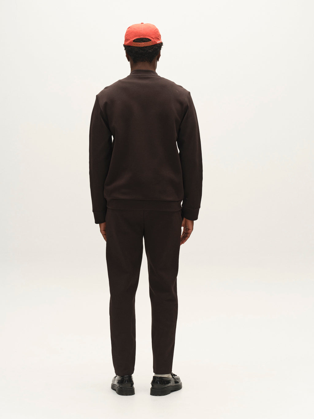 A person stands facing away, wearing the Gabba GABnik Camp Lock Sweat in Meteorite—a dark brown tracksuit crafted from mid-weight interlock fabric—paired with black shoes and a red cap, set against a plain white background.