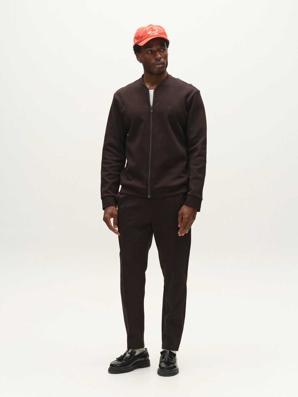 A man stands against a plain background, wearing Gabba’s GABmonza Cropped Tasca Lock Pant in Meteorite—a relaxed, straight-fit style crafted from mid-weight interlock fabric—paired with a dark brown zip-up jacket, black shoes, and a red cap. He is looking slightly to the side with his hands relaxed by his sides.