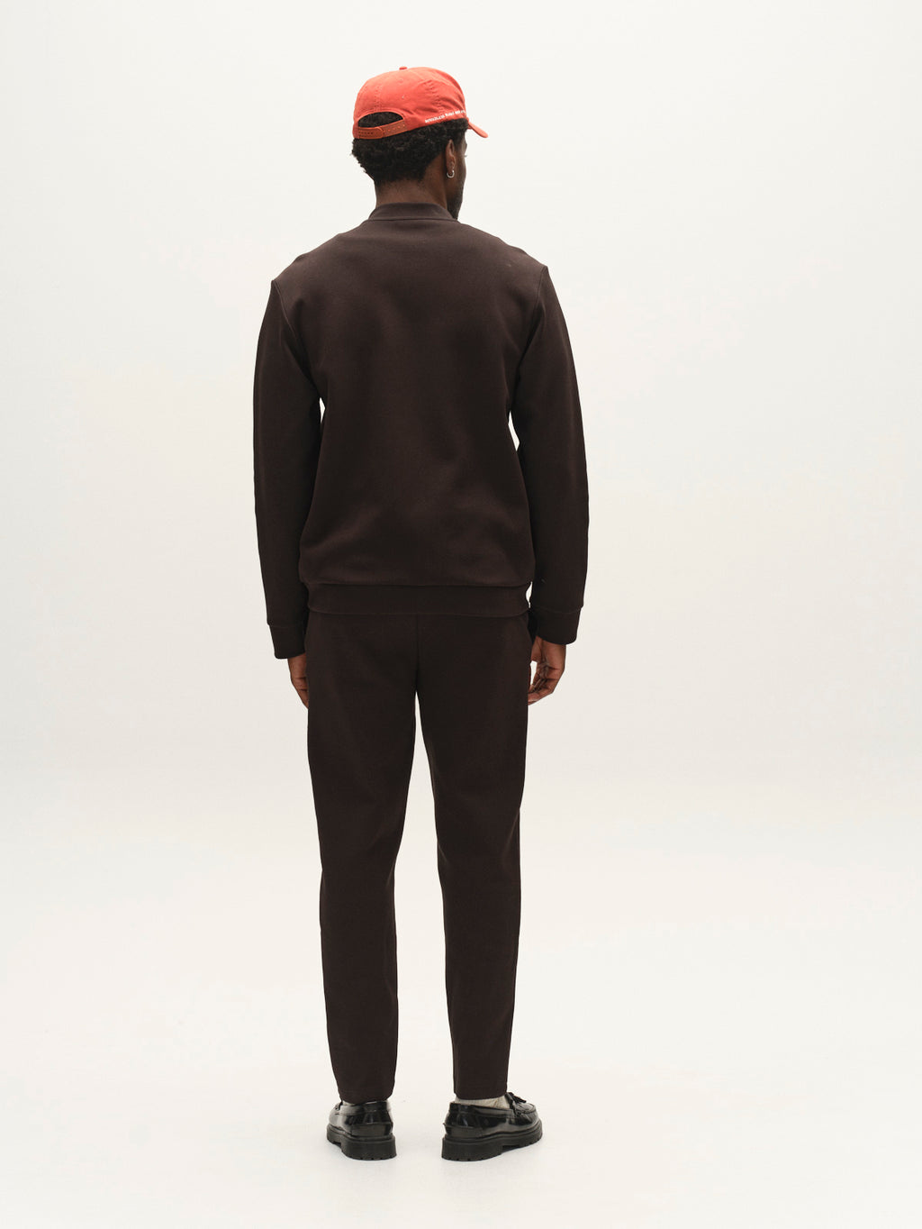 A person stands facing away from the camera, wearing a black outfit featuring the Gabba GABmonza Cropped Tasca Lock Pant in Meteorite with matching shoes and a red cap against a plain, light background.