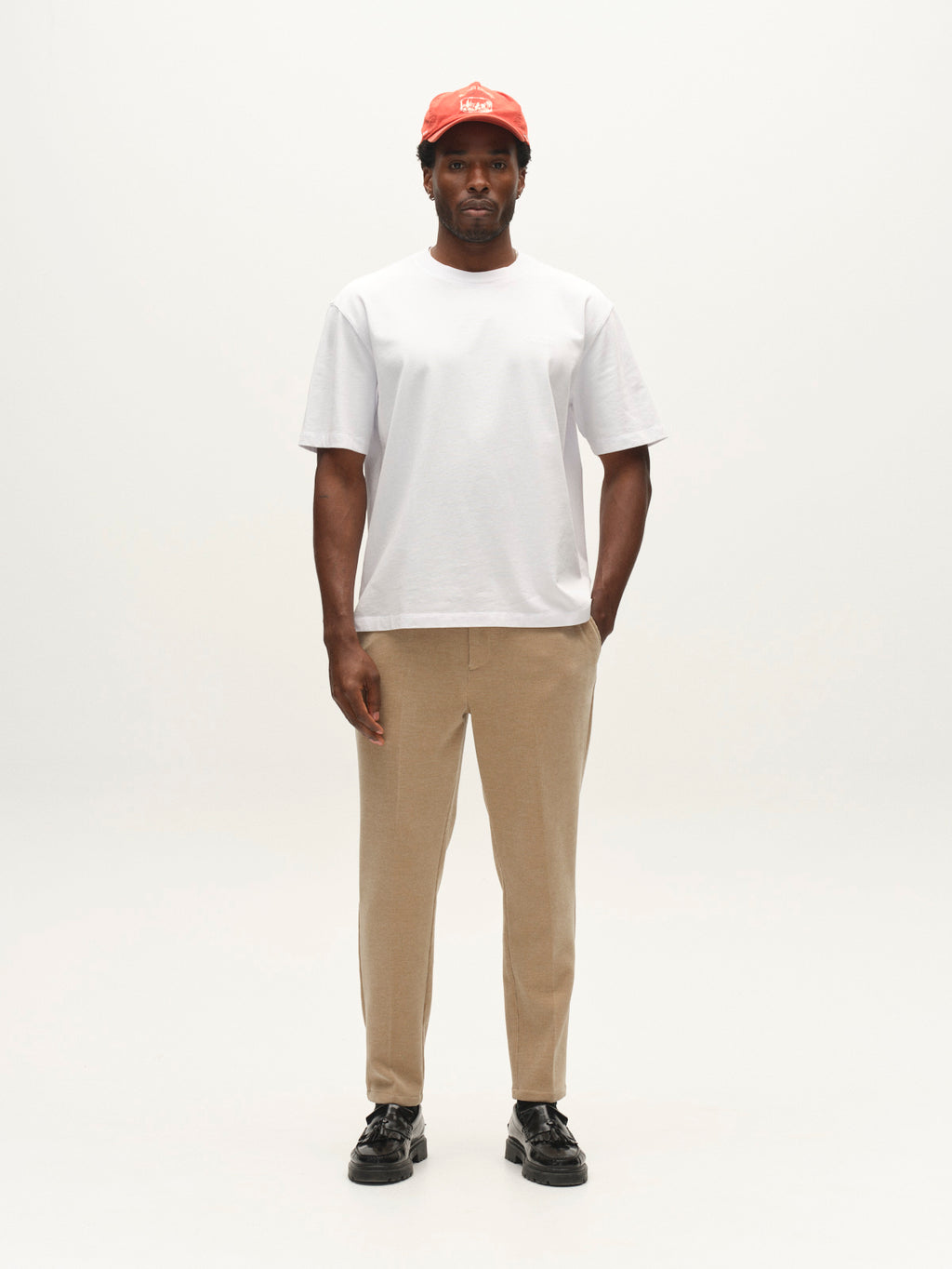 A man stands facing forward, wearing a red cap, white T-shirt, Gabba GABmonza Cropped Tasca Fune Pant in Sand Crocker, and black shoes, against a plain white background.