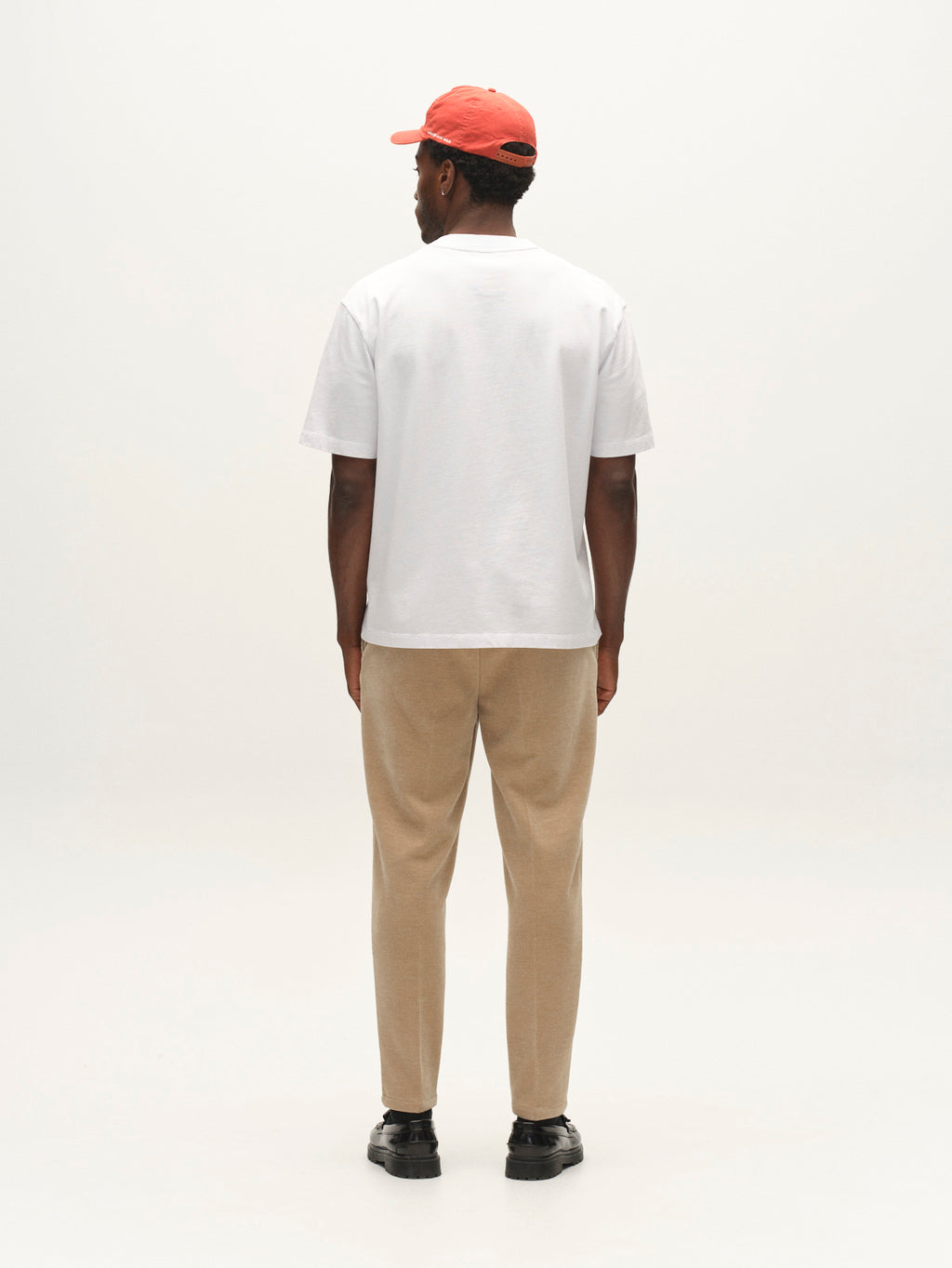 A person stands facing away, wearing a red cap, white t-shirt, Gabba GABmonza Cropped Tasca Fune Pant in Sand Crocker, and black shoes, against a plain light background.
