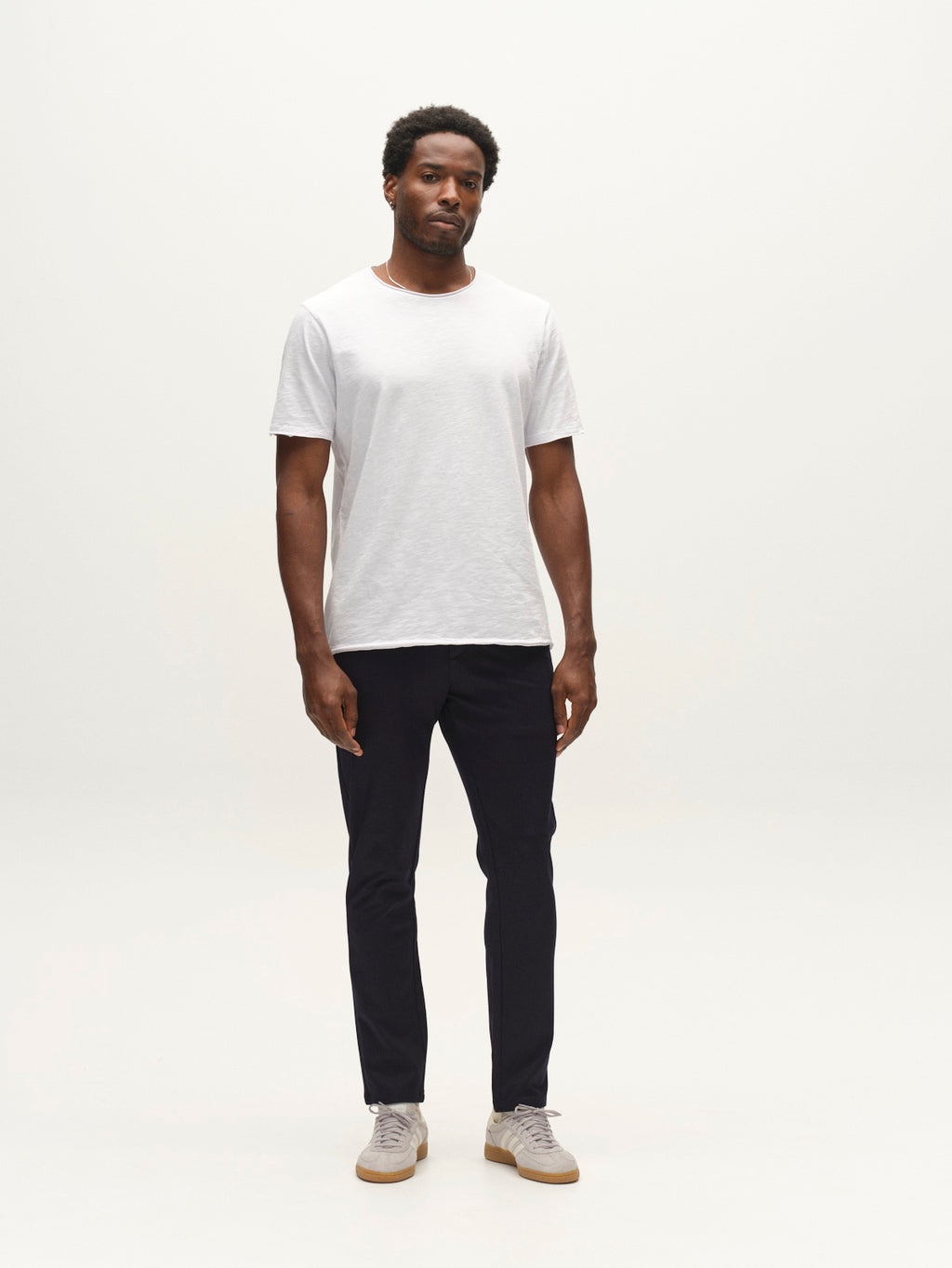 A man stands against a plain white background, wearing a light gray T-shirt, the Gabba GABpaul Dotty Pant in navy—a slim-fit chino—and light-colored sneakers. He faces forward with a neutral expression and relaxed posture.
