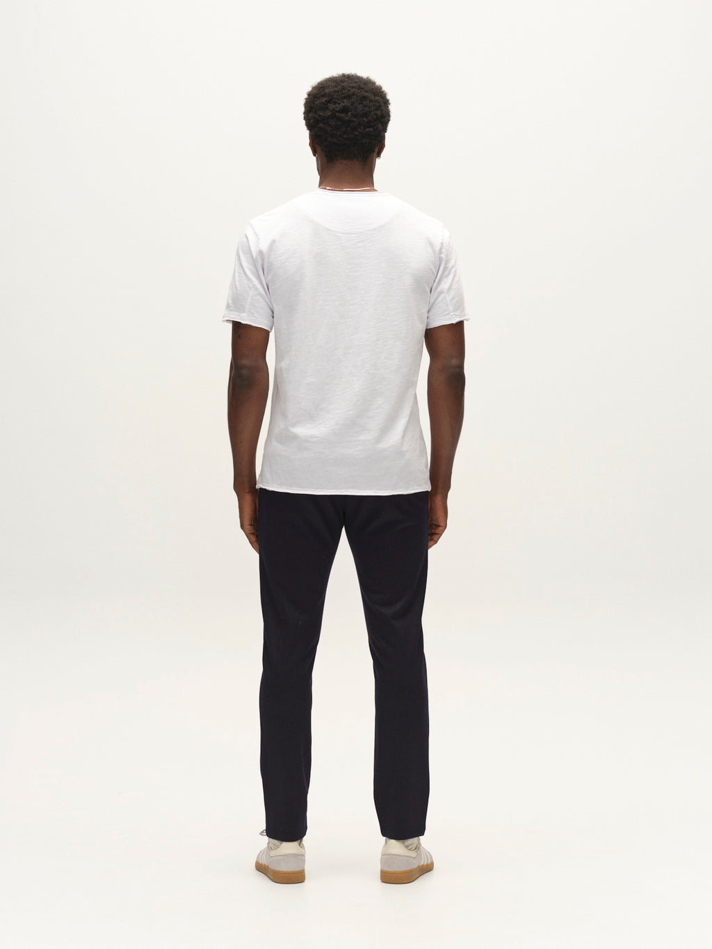 A man stands facing away from the camera, wearing a plain white t-shirt, light-colored shoes, and Gabba’s GABpaul Dotty Pant in navy—a pair of dark slim chino fit pants crafted from superstretch fabric—against a white background.