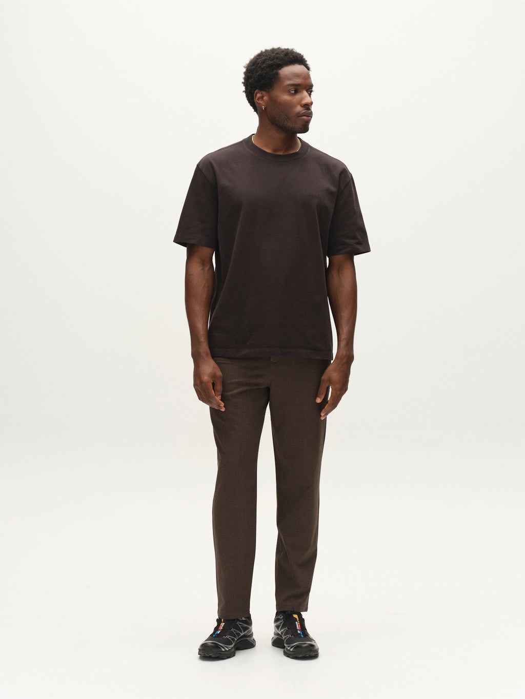 A man stands against a plain white background, wearing a black t-shirt, black sneakers, and Gabba GABjoe-pas Rubin Pant in Brown Slate. He looks slightly to his left with his arms relaxed by his sides.
