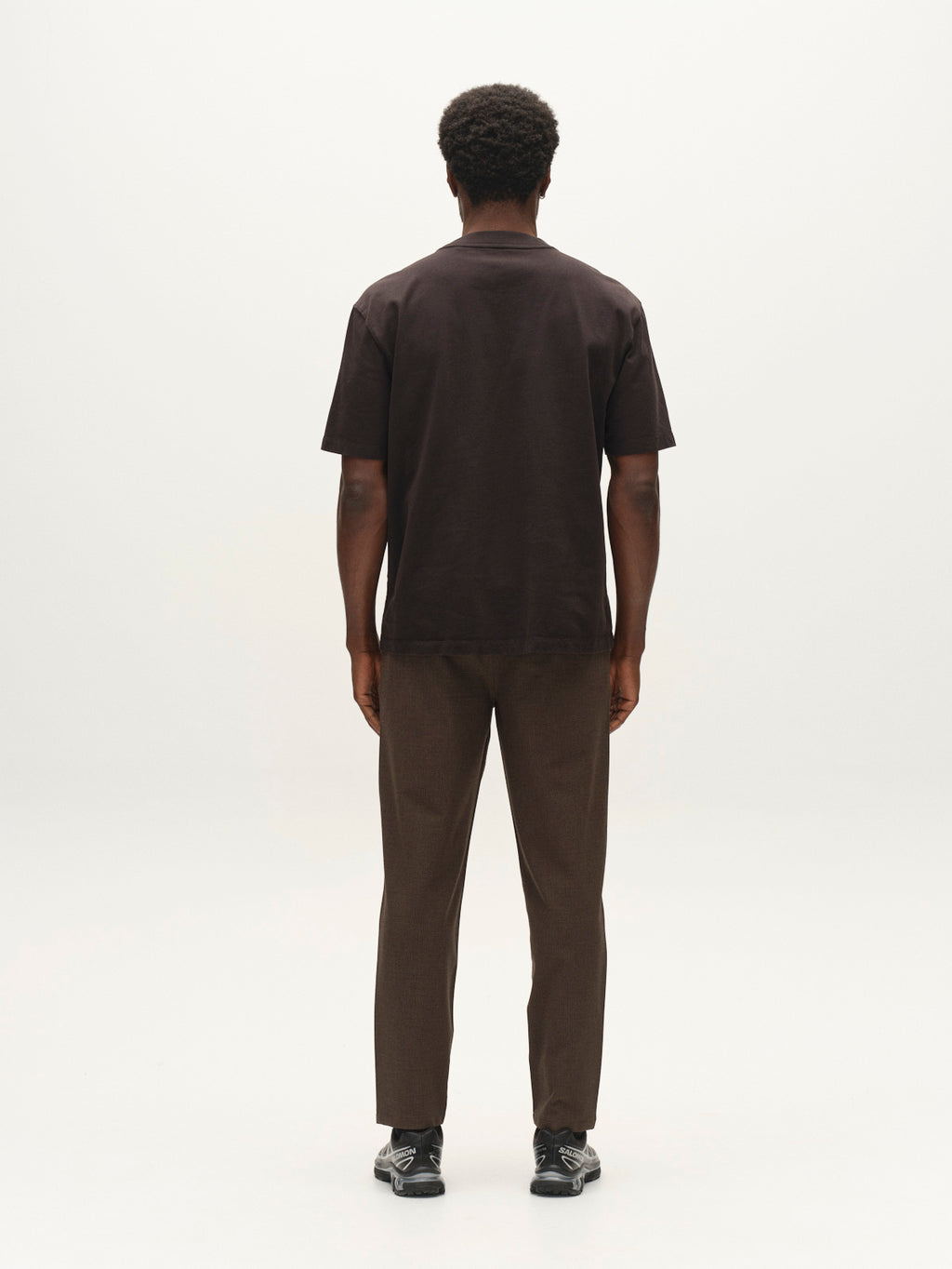 A man stands facing away from the camera, wearing a dark short-sleeved shirt, black shoes, and Gabba’s GABjoe-pas Rubin Pant in Brown Slate, all set against a plain white background.