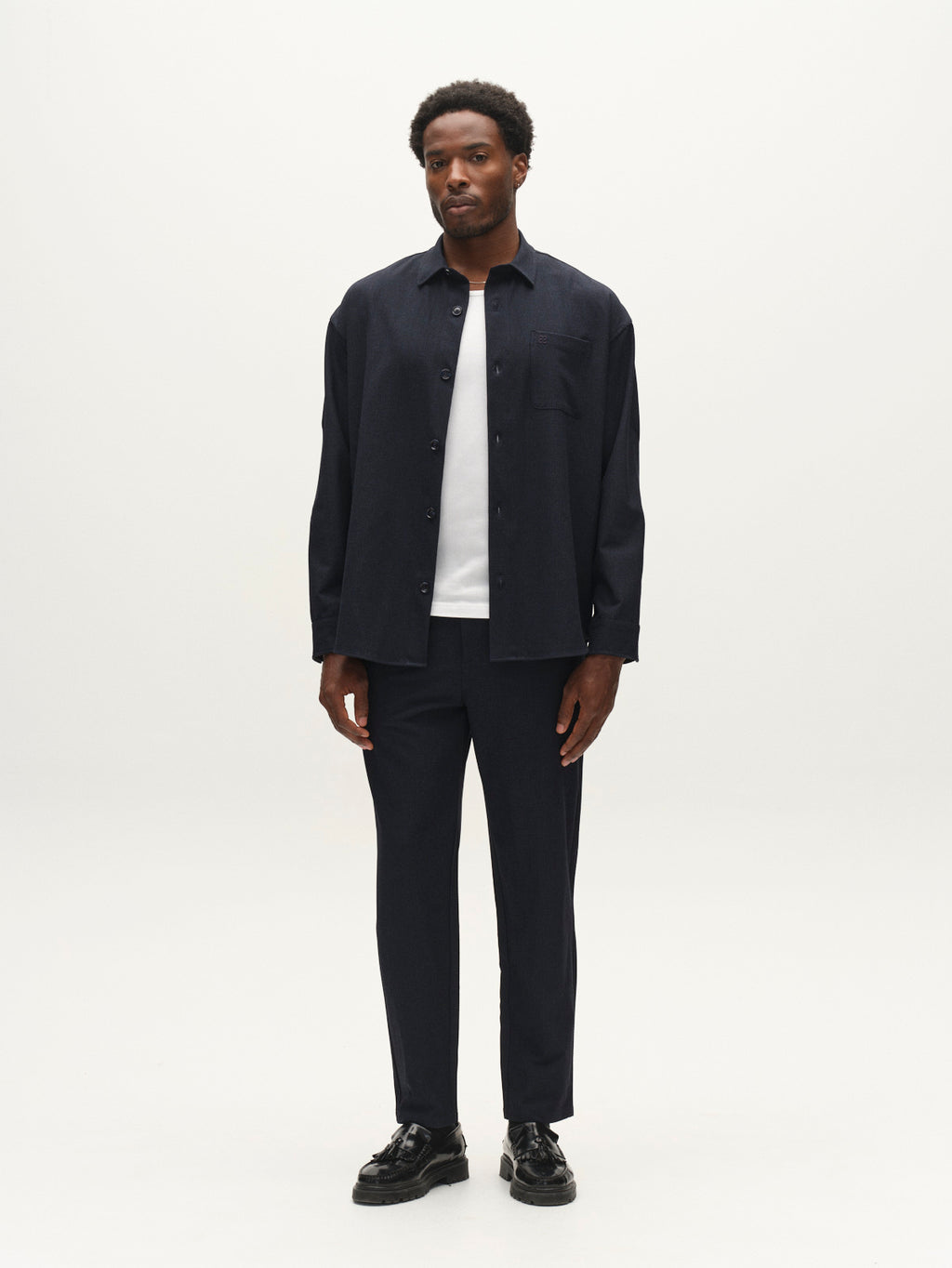 A man stands against a plain white background, wearing a white T-shirt, a dark navy button-up shirt, and Gabba’s GABjoe-pas Rubin Pant in Blue Black—a pair of tapered fit pants crafted from stretch dobby fabric—paired with black dress shoes. He has short hair and a neutral expression.