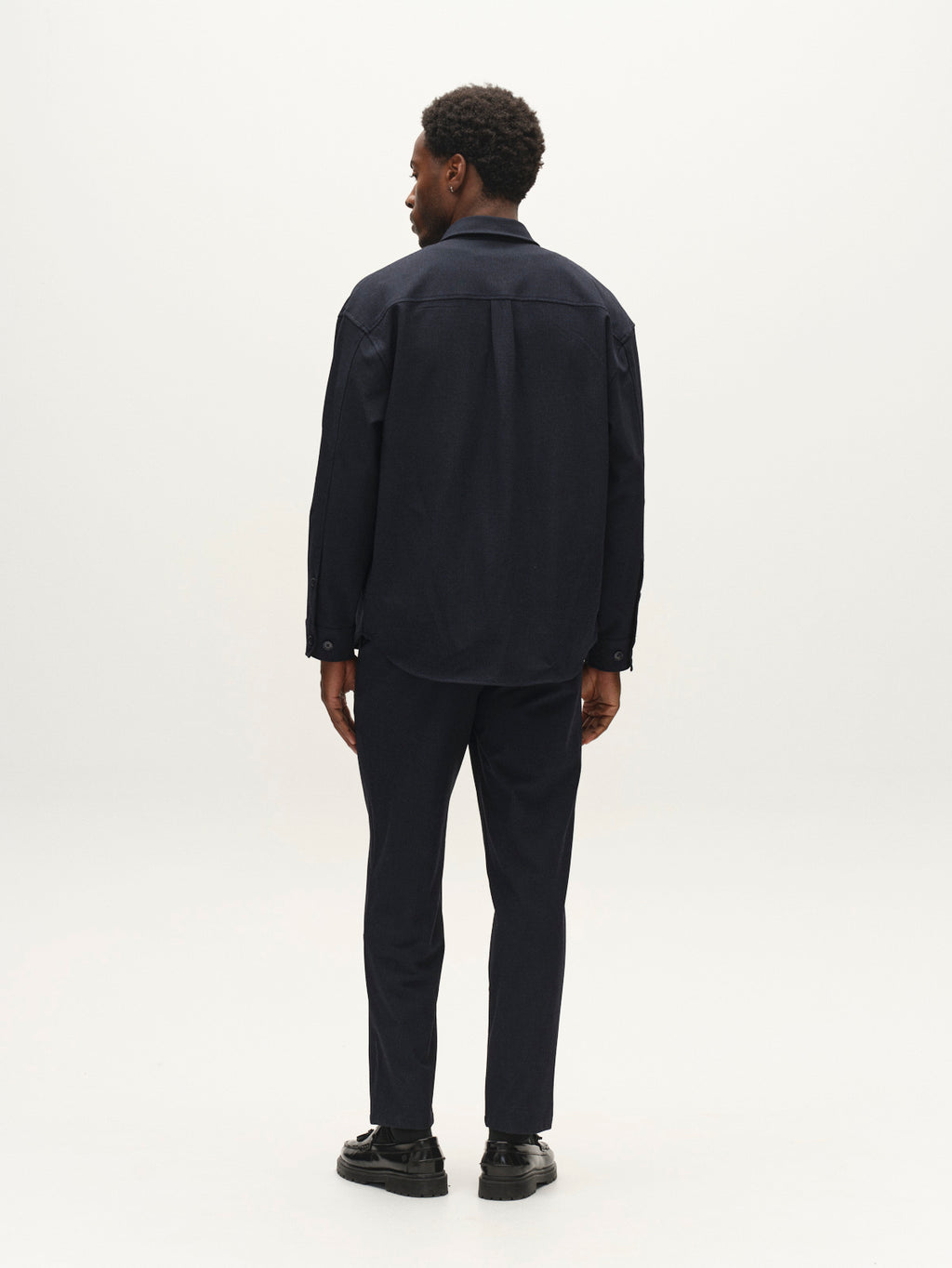 A man stands facing away, wearing a dark navy shirt, Gabba’s GABjoe-pas Rubin Pant in Blue Black—tailored from stretch dobby fabric—and black shoes against a plain, light background.