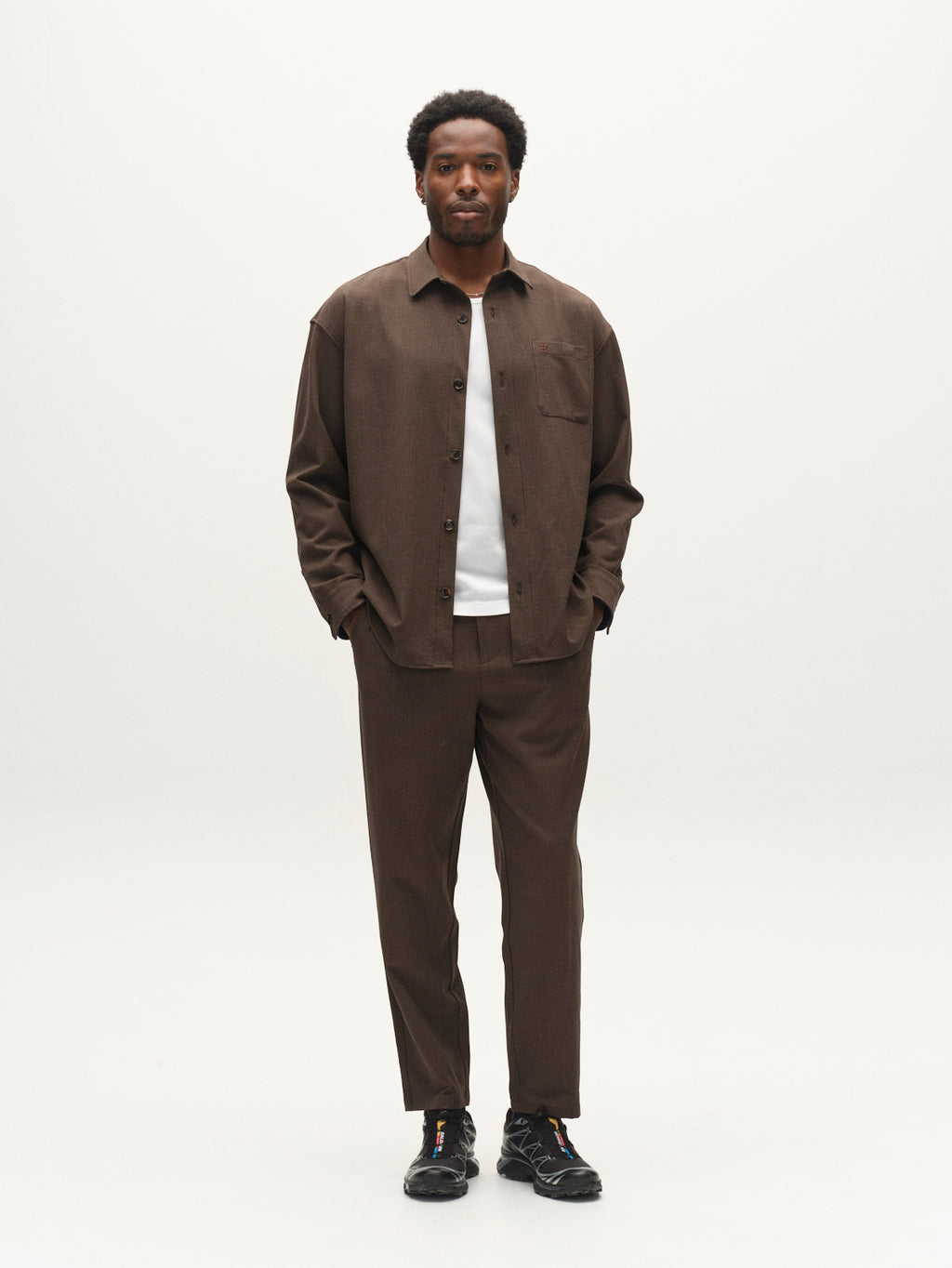A man stands facing forward, wearing the Gabba GABdavid Rubin Pant in Brown Slate, styled with a matching brown button-up shirt, a white t-shirt underneath, and black sneakers. The pants feature a loose tapered fit crafted from stretch dobby fabric. The background is plain and light-colored.