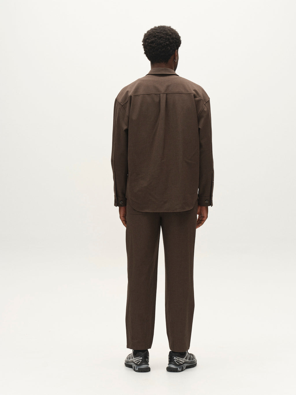 A person stands facing away, dressed in a brown long-sleeve shirt and Gabba GABdavid Rubin Pant in Brown Slate, featuring a loose tapered fit, paired with black sneakers, set against a plain white background.