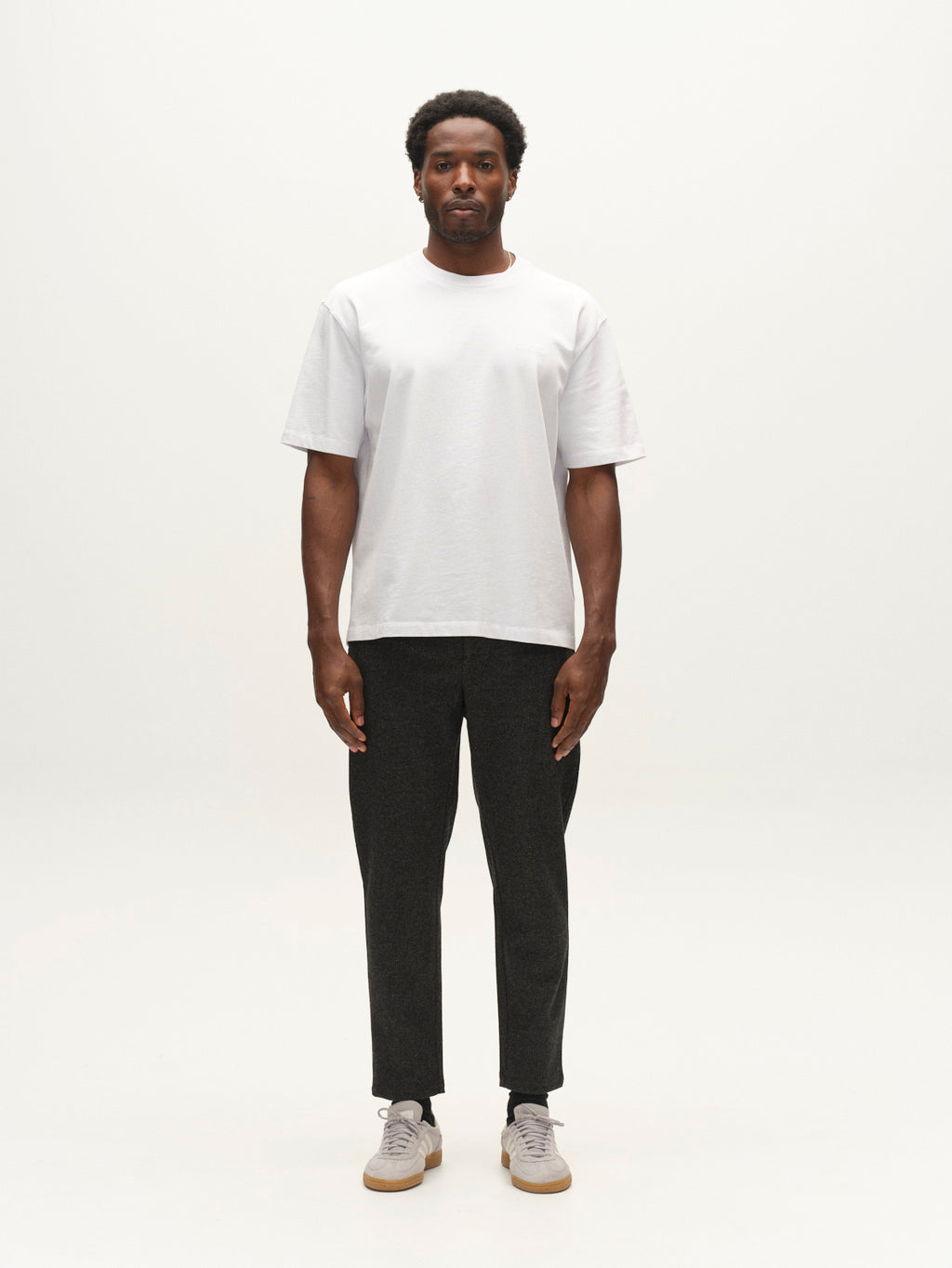 A man stands facing forward against a plain white background, wearing a white T-shirt, light gray sneakers, and Gabba’s GABmonza Cropped Tasca Zaffe GRS Pant in dark grey—a relaxed straight fit style featuring an elastic waistband and wool-look finish.
