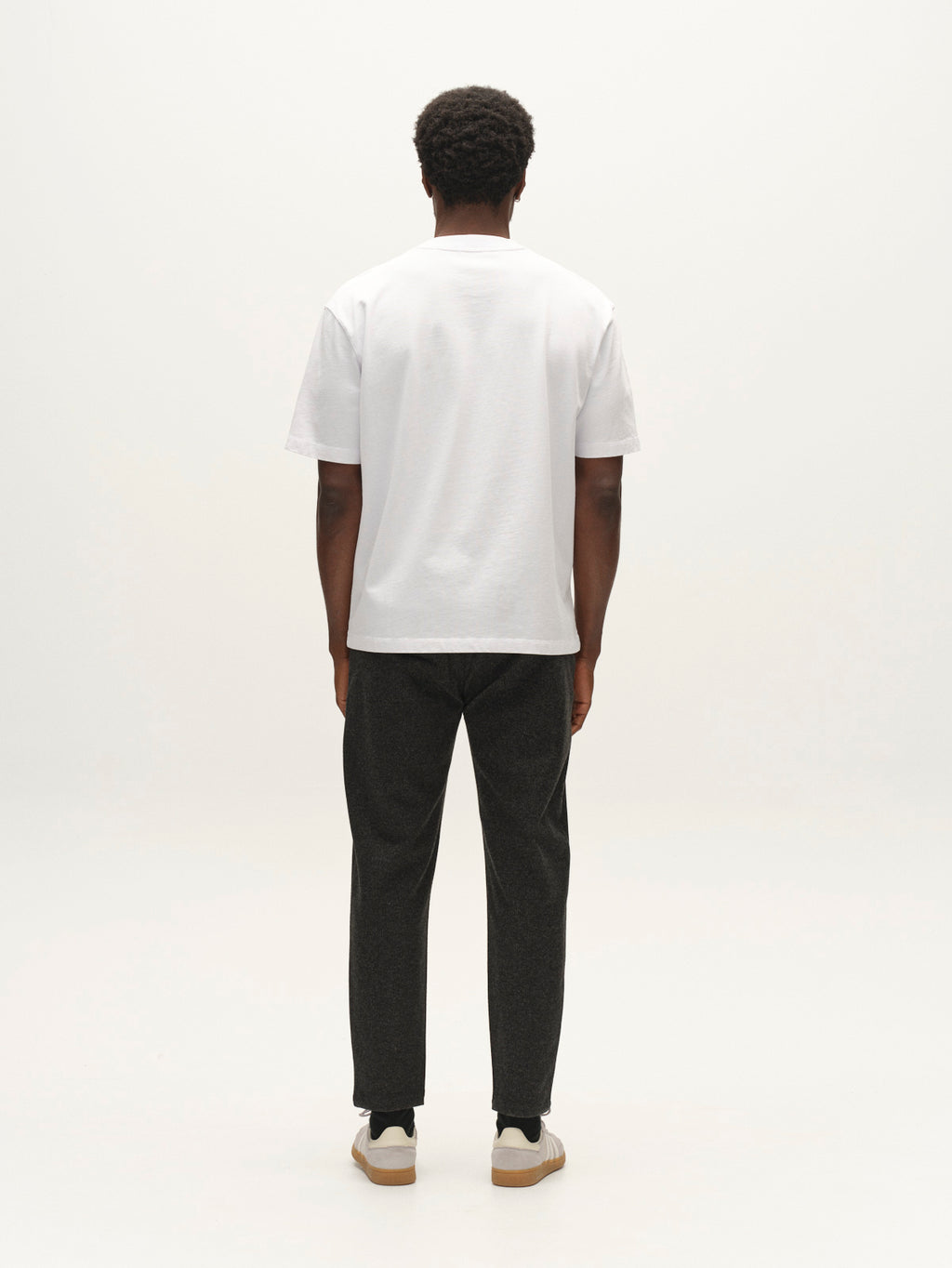 A person with short curly hair, wearing a plain white T-shirt, Gabba GABmonza Cropped Tasca Zaffe GRS Pant in dark grey featuring a relaxed straight fit and an elastic waistband, and sneakers, stands facing away from the camera against a plain light background.