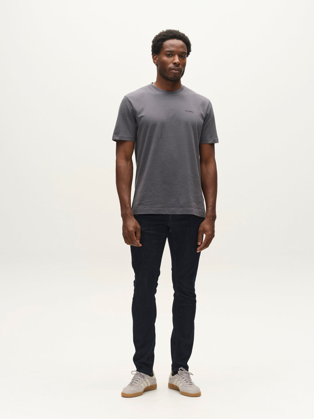 A man stands facing forward against a plain white background, wearing a gray t-shirt, light-colored sneakers, and Gabba GABjones Vanew Rinse Jeans in dark blue denim.