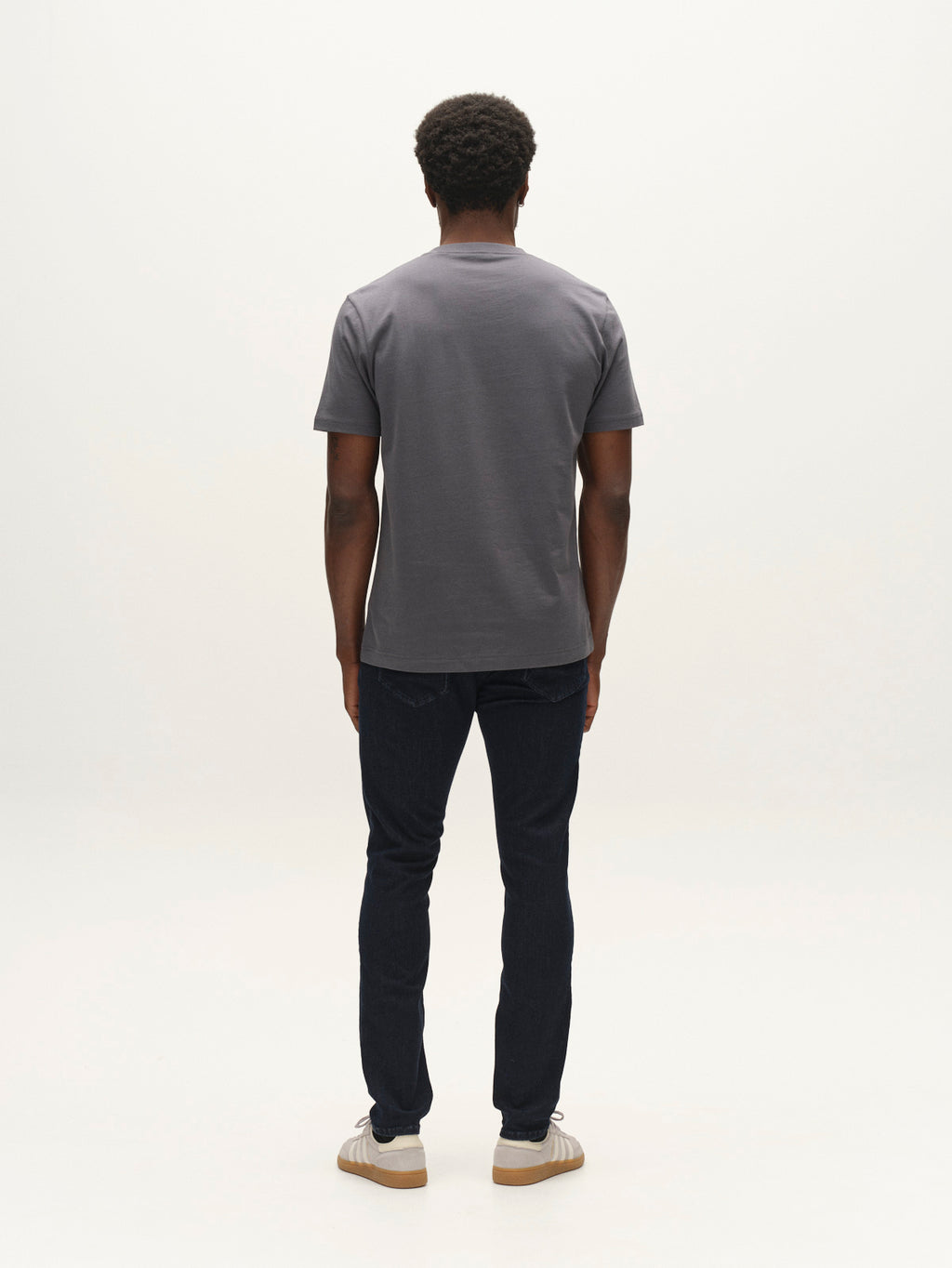 A man with short curly hair stands facing away, wearing a gray t-shirt, Gabba GABjones Vanew Rinse Jeans in dark blue denim, and white sneakers, against a plain white background.