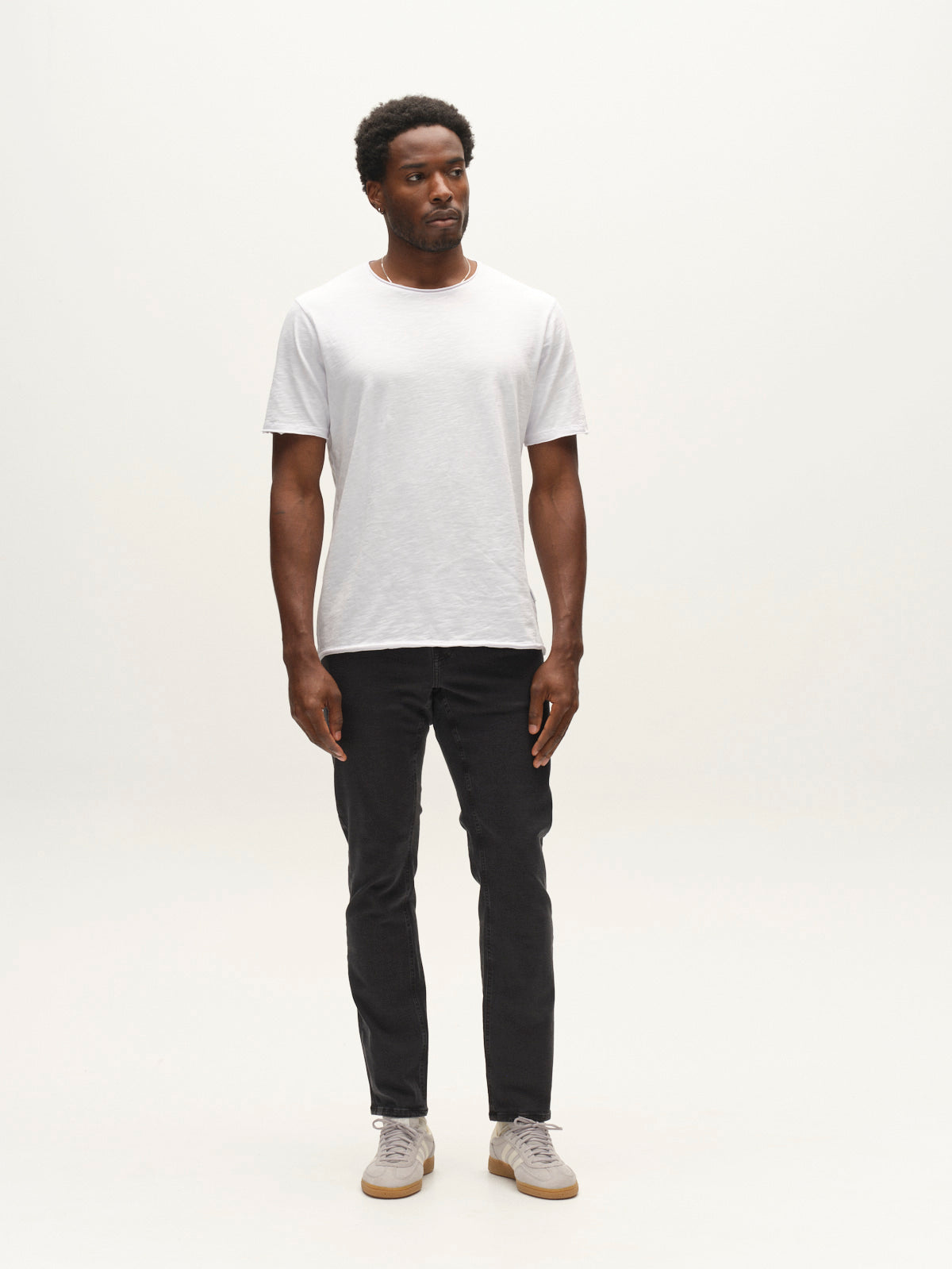 A man stands against a plain white background wearing a white t-shirt, light-colored sneakers, and Gabba GABjones Hesky Jeans in black denim. He looks slightly to the side with a neutral expression and his arms relaxed by his sides.