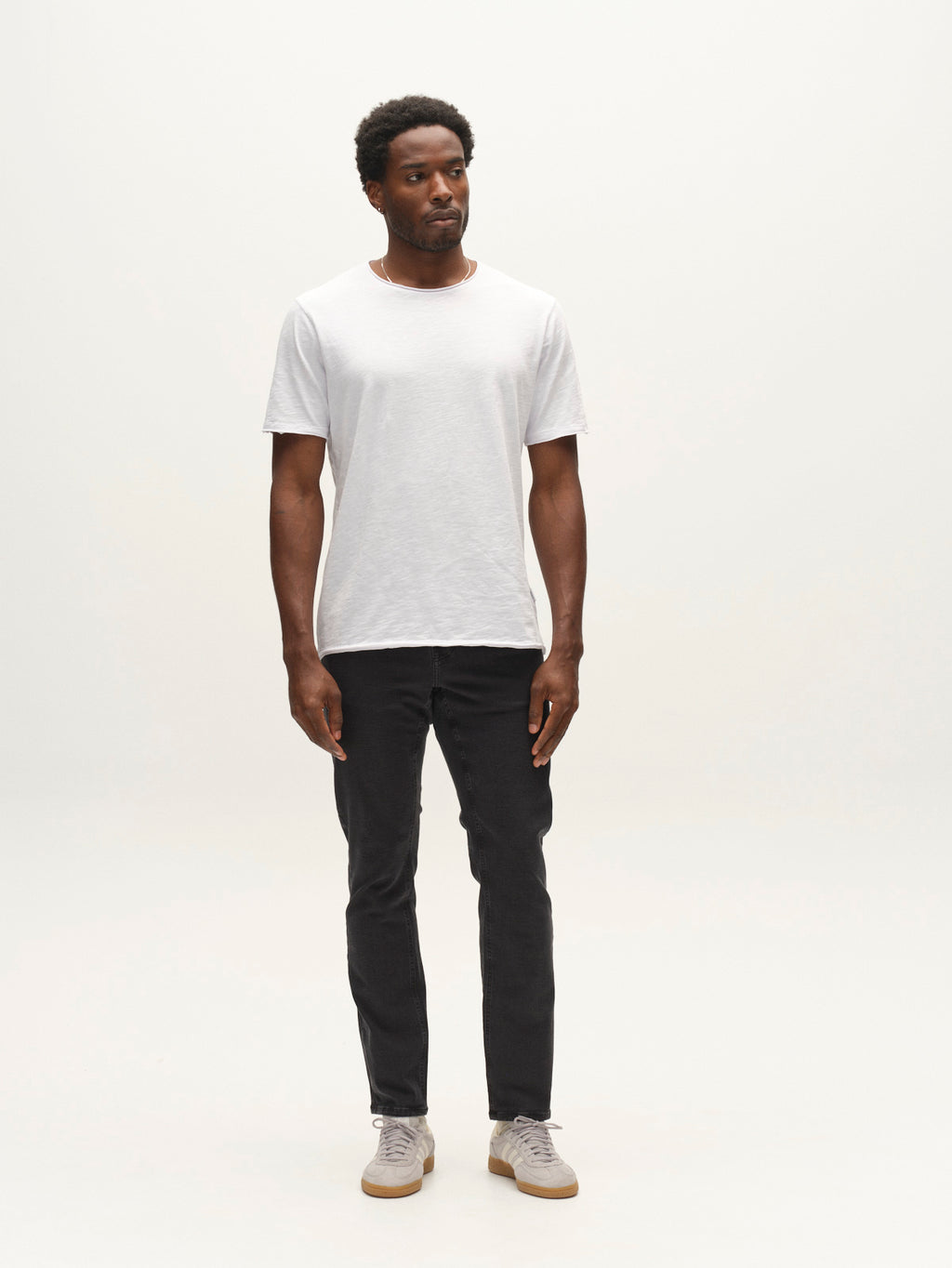 A man stands against a plain white background wearing a white t-shirt, light-colored sneakers, and Gabba GABjones Hesky Jeans in black denim. He looks slightly to the side with a neutral expression and his arms relaxed by his sides.