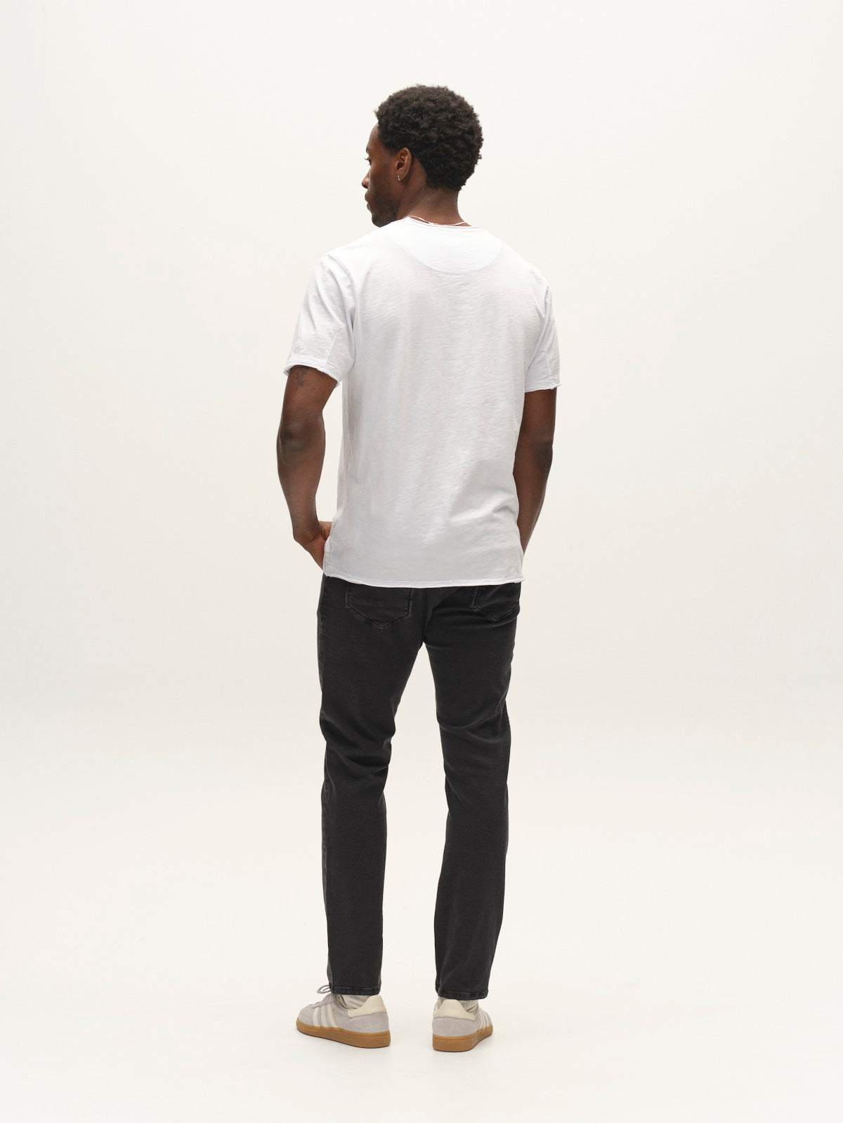 A man stands facing away against a plain light background, wearing a plain white t-shirt, white sneakers, and Gabba GABjones Hesky Jeans in black denim.