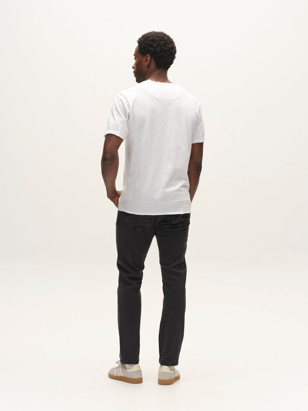 A man stands facing away against a plain light background, wearing a plain white t-shirt, white sneakers, and Gabba GABjones Hesky Jeans in black denim.