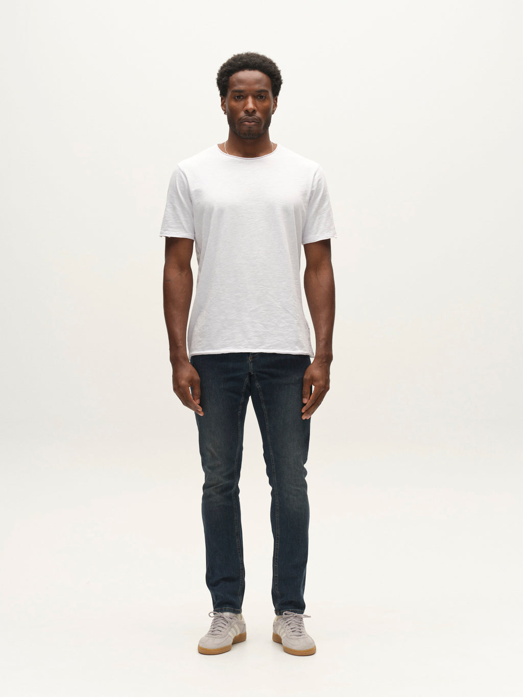 A man stands facing forward, wearing a plain white t-shirt, Gabba GABjones2 Nham RS2113 Jeans in dark blue denim crafted from performance stretch fabric, and gray sneakers, against a simple white background.
